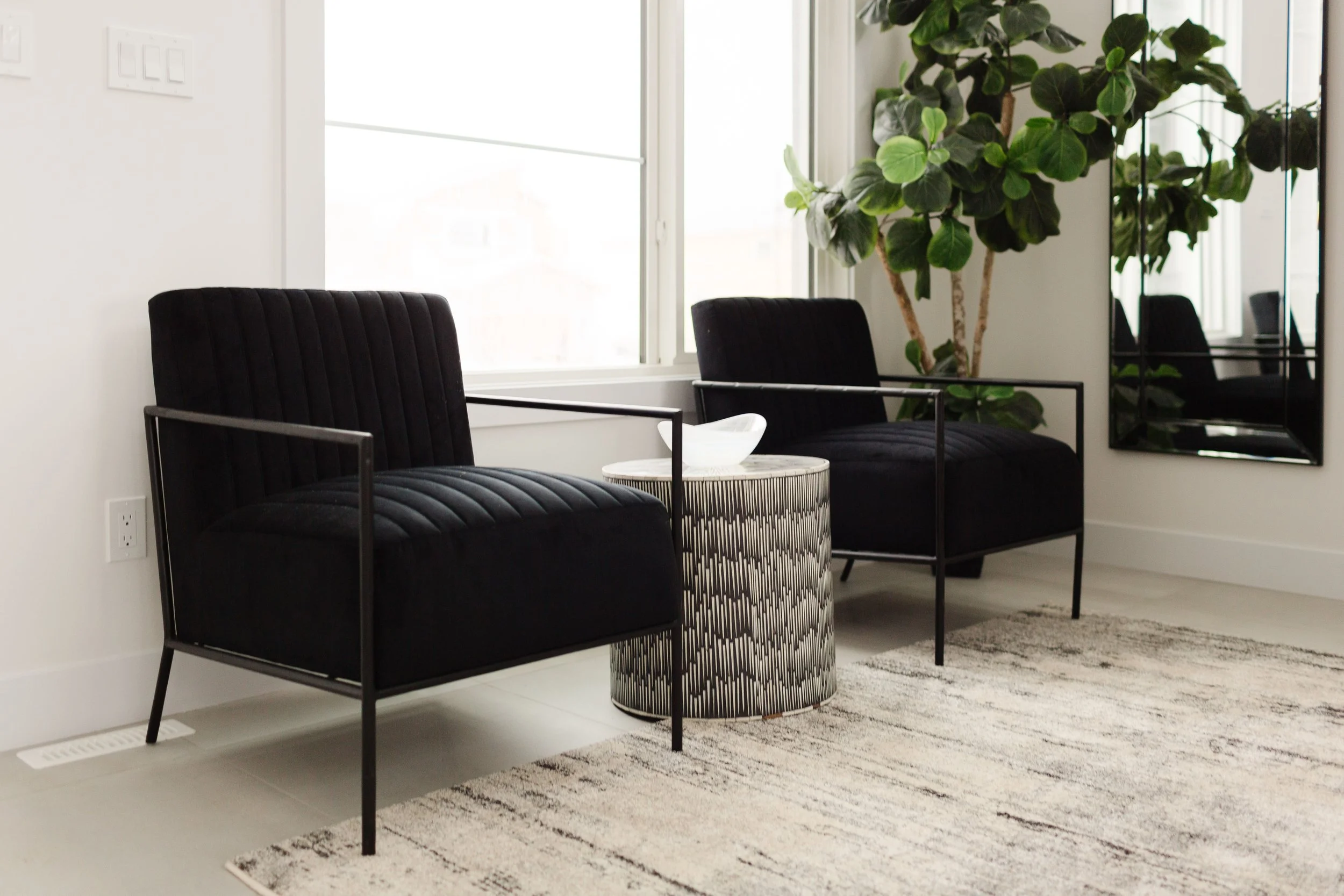 Photo of two black armchairs next to an elegant black and white side table and rugs with natural lighting shining in the window behind