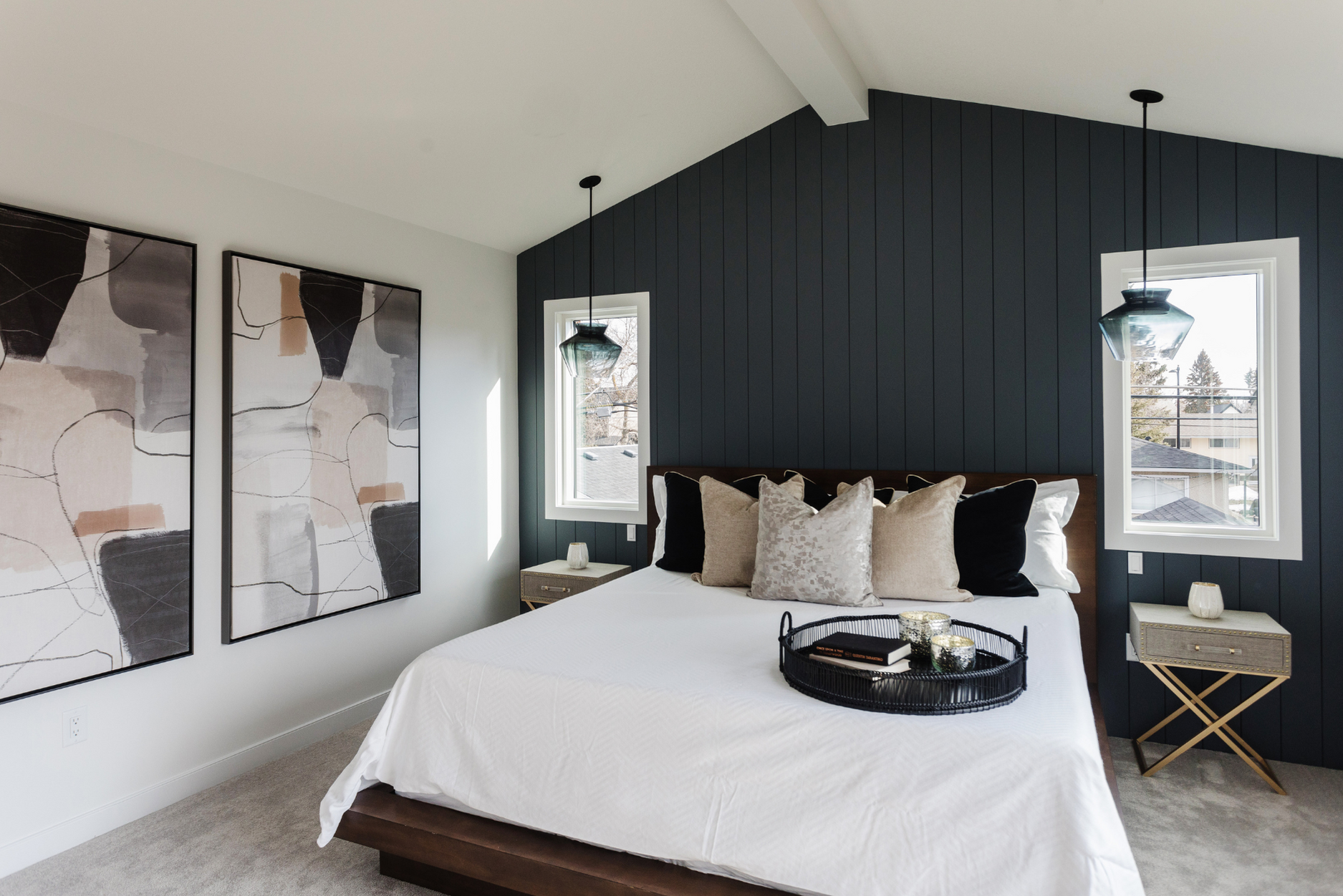 Photo of a bedroom with white king bed with throw pillow and tray, a blue wood panel accent wall, and windows behind with nighstands on either side of the bed
