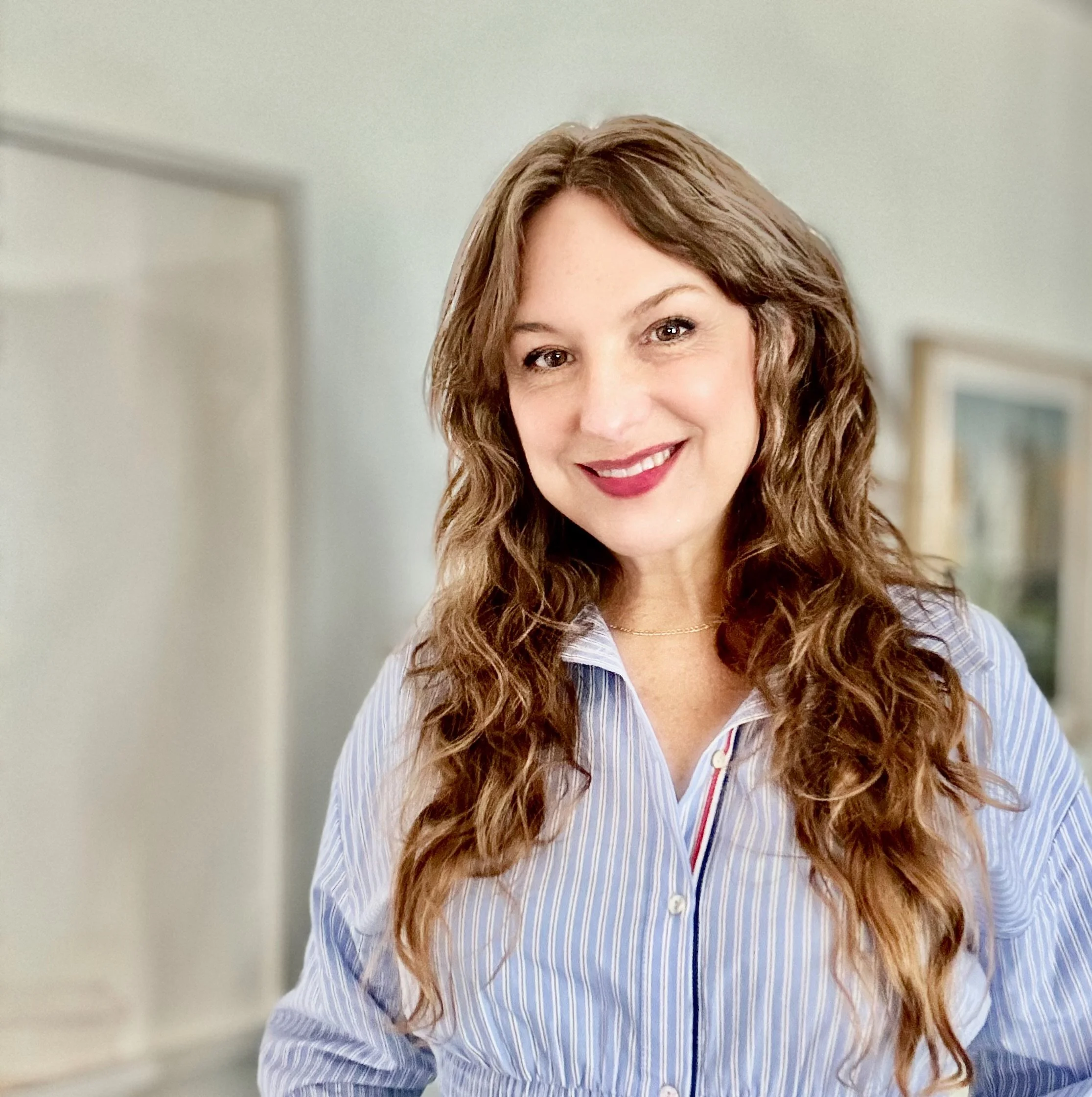 Amber Noland. A woman with long curly brown hair smiling, wearing a blue and white striped shirt, standing indoors with framed pictures on the wall in the background.