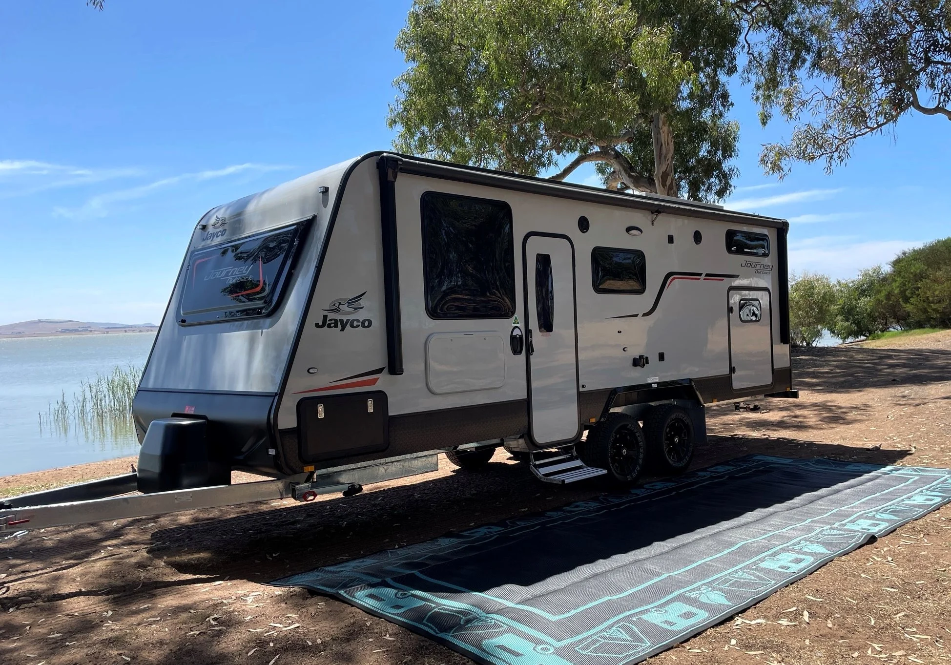 A silver and black Jayco Journey travel trailer parked on a blanket near a lakeshore with trees and clear blue sky.