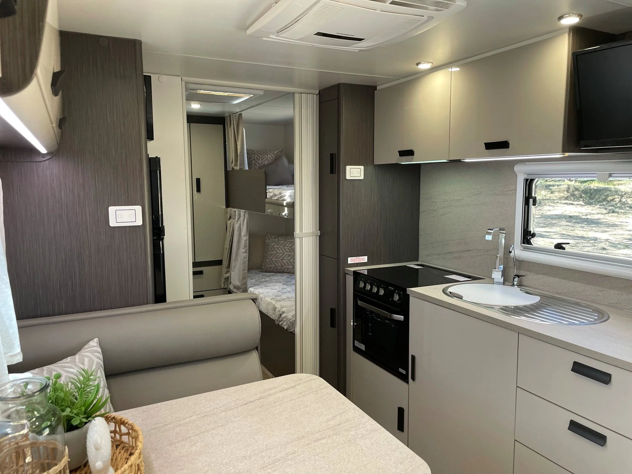 Interior of a camper or RV kitchen and living area with a small sink, stove, cabinets, a television, and a bed area with bunk beds in the background.