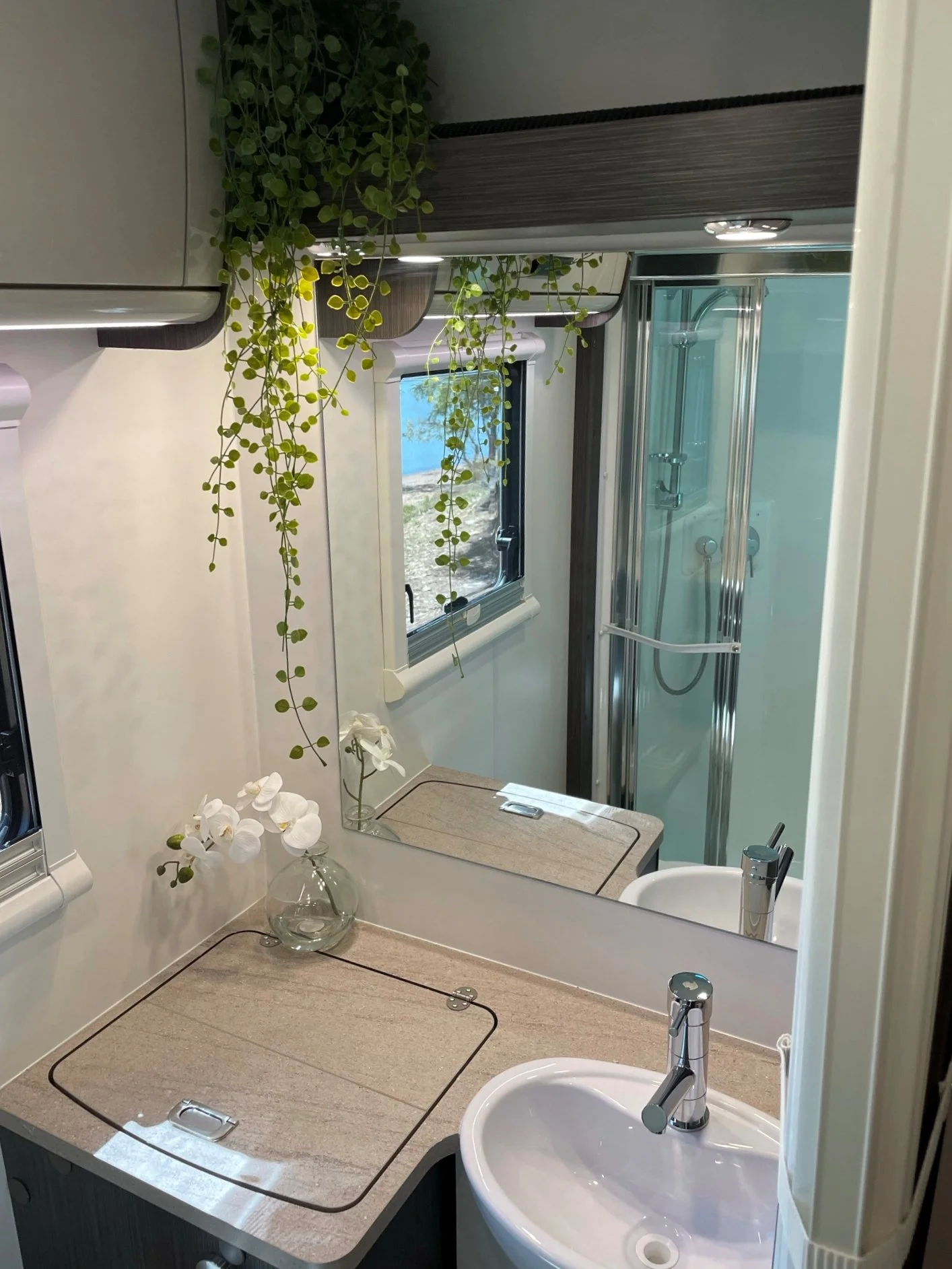 Bathroom with a beige countertop, a round white sink with a modern faucet, a mirror, a small window, a shower with clear glass doors, and a hanging green plant and white flowers in a glass vase.