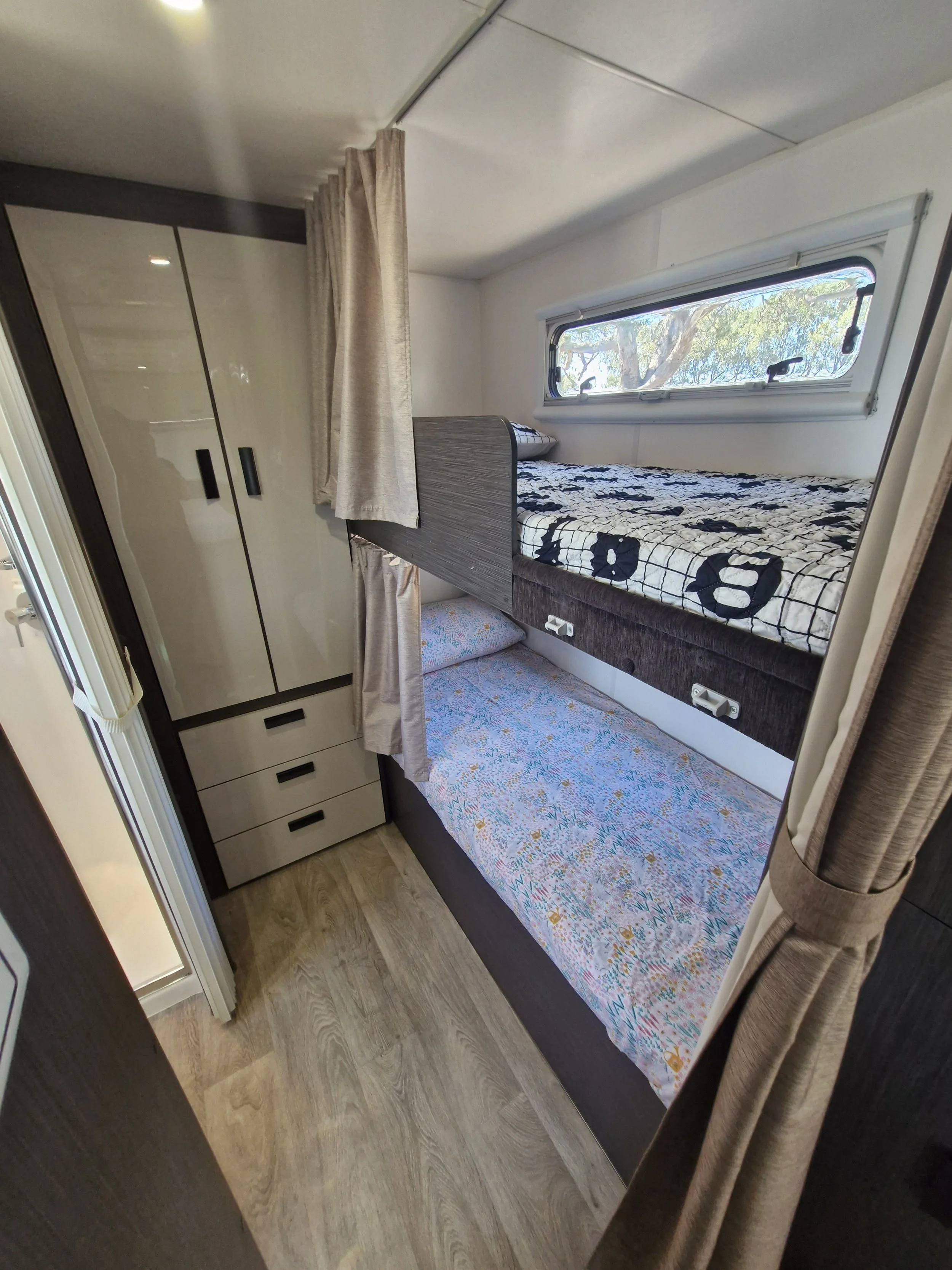 Interior of a compact camper trailer with bunk beds, wardrobe, and curtains.