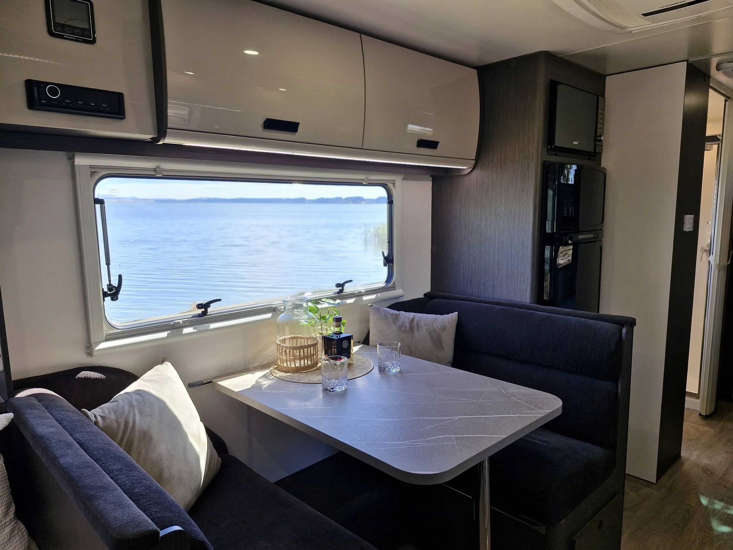 A cozy interior of a boat or RV featuring a table by a large window overlooking a body of water, with seating and modern cabinetry.