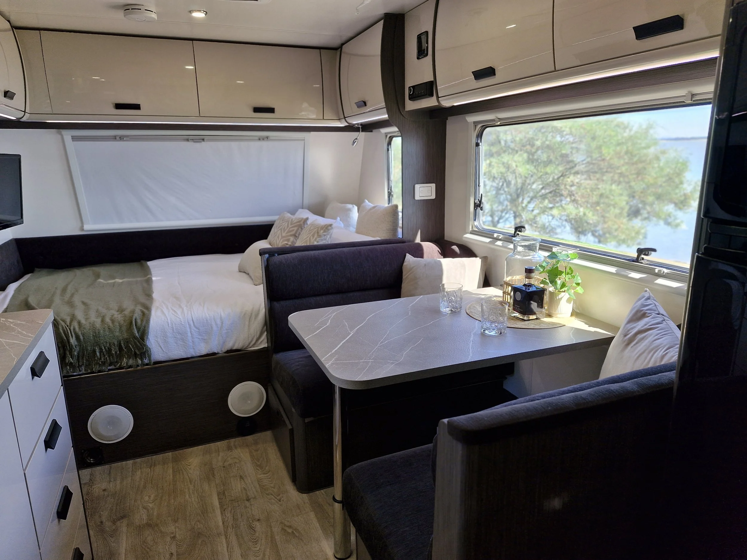 Interior of a compact camper or RV with a bed, small dining area, and large window showing a tree outside.