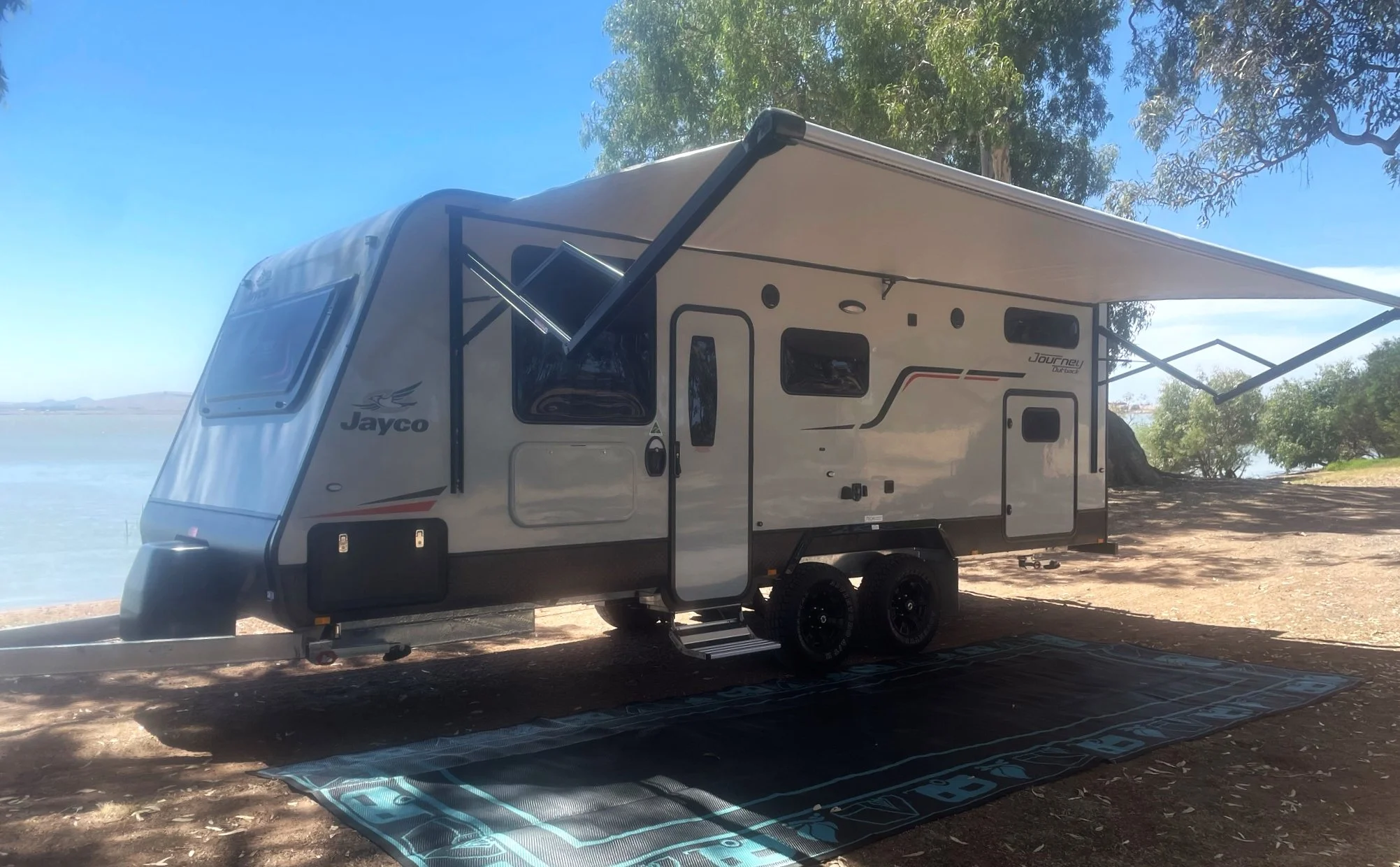 A white Jayco travel trailer with a black and red design, parked on a dirt area near water and trees, with an extended awning and a black mat on the ground.
