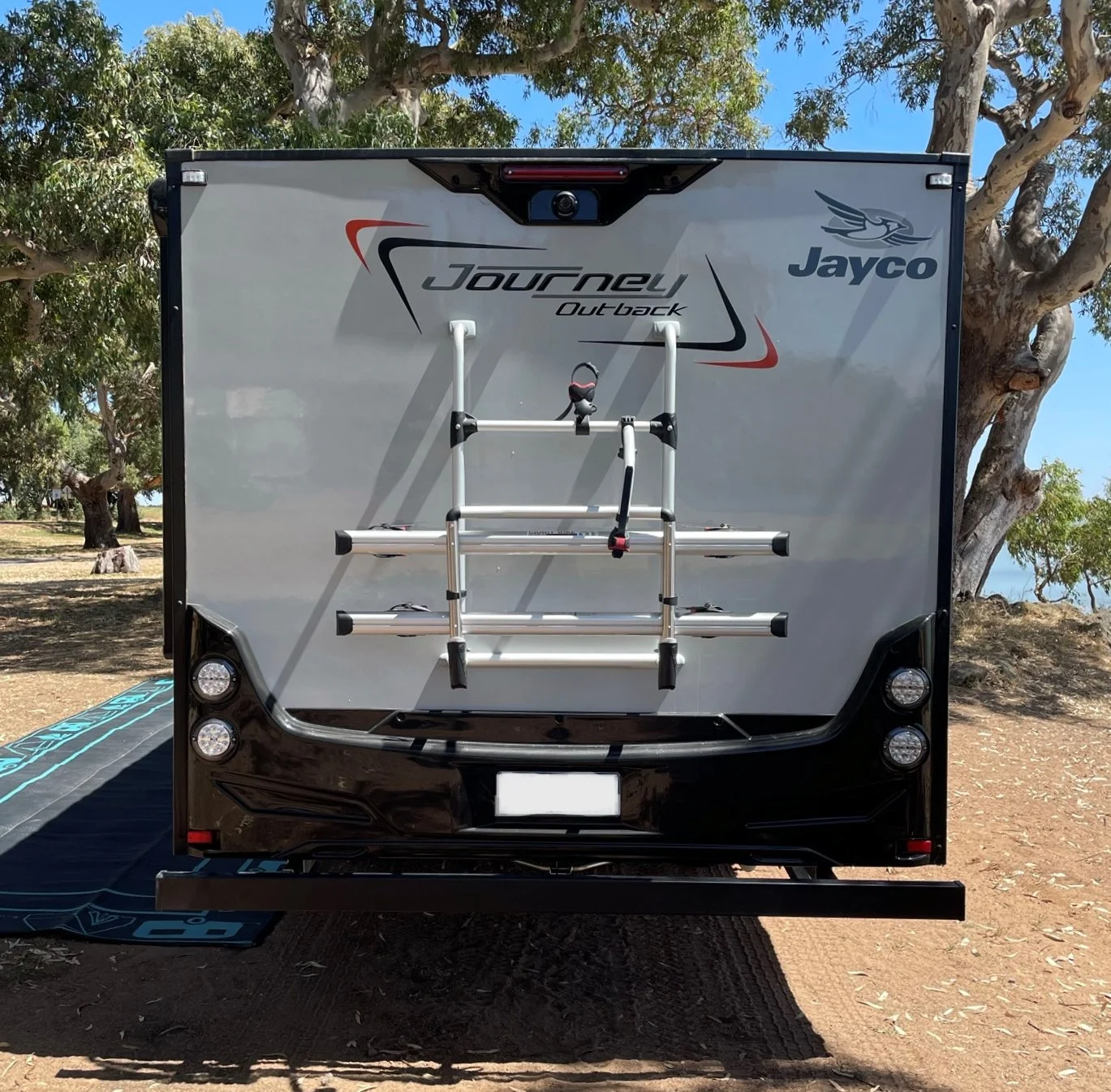 Rear view of a Jayco Journey Outback RV with a bike rack mounted on the back. The RV is parked on a dirt surface with trees and blue sky in the background.