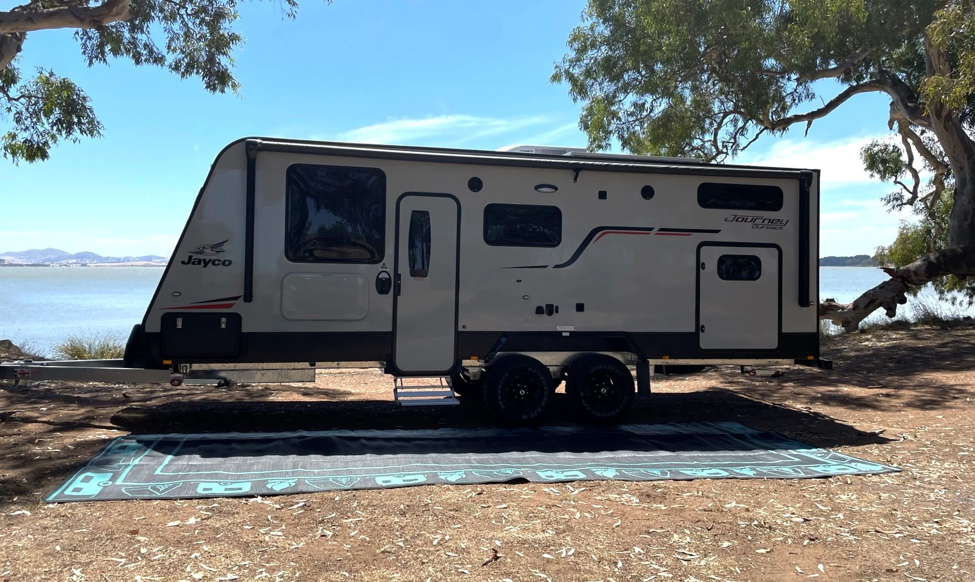 Travel trailer parked on a dirt area near a body of water with trees and mountains in the background.