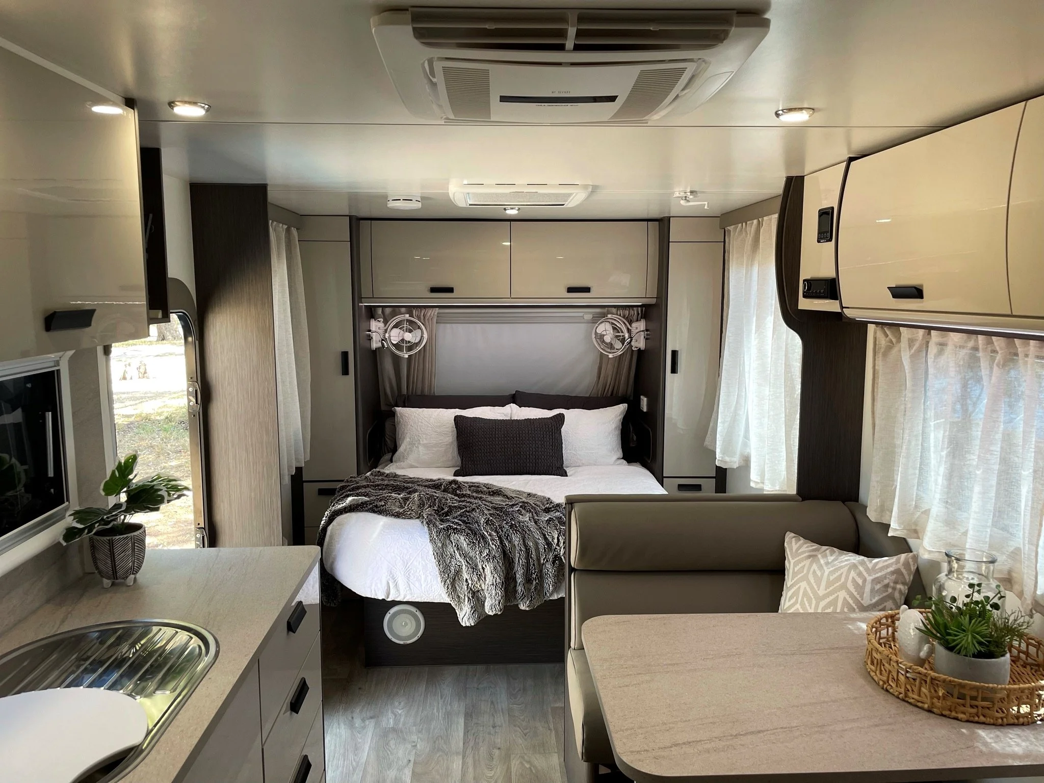 Inside of a modern RV or camper, featuring a bed with pillows, small fans on either side of the headboard, a dining area with a table, a couch with a decorative pillow, and a kitchenette with a sink, cabinets, and a window with curtains.