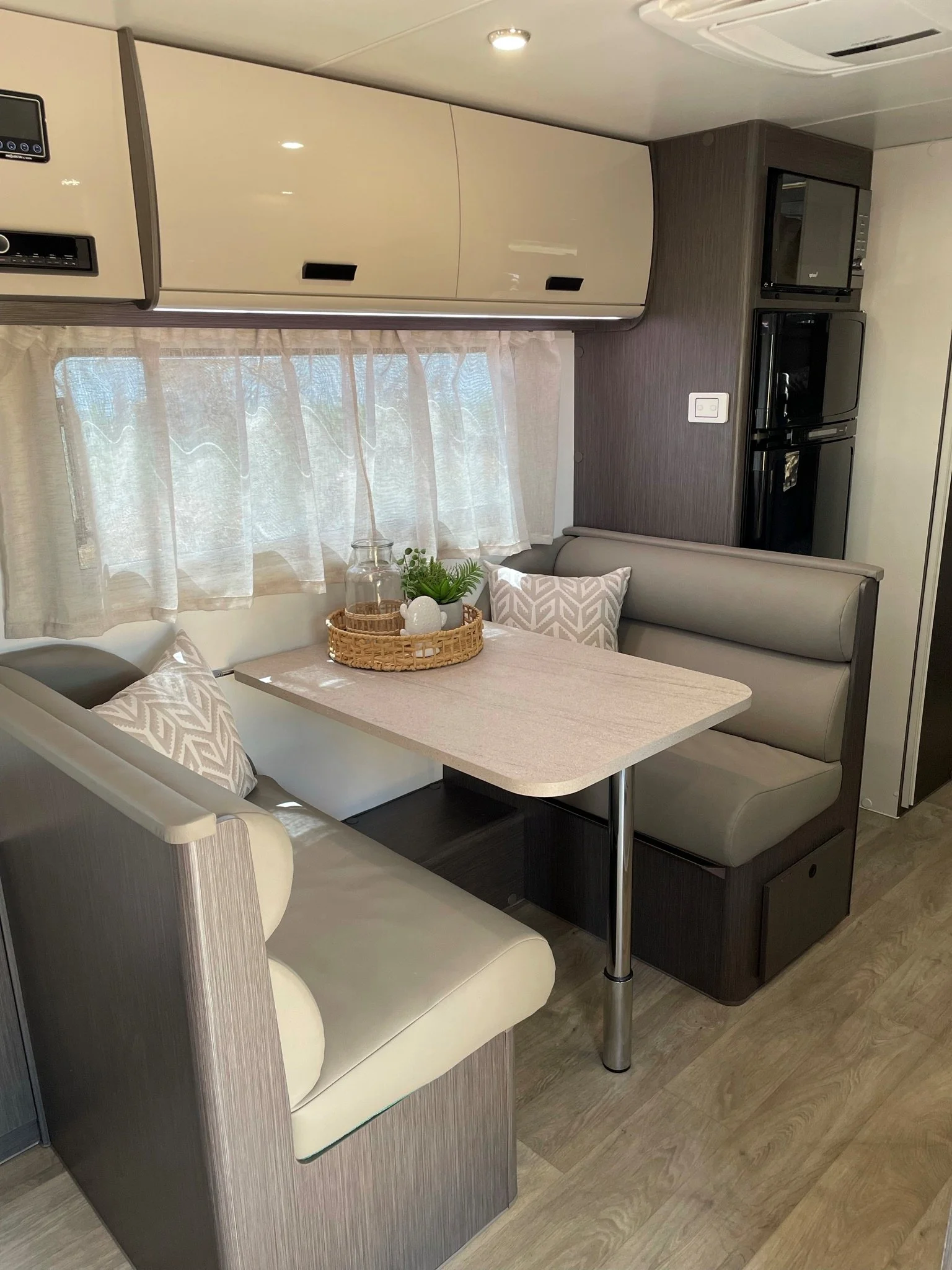 A cozy dinette area inside a camper or RV, featuring beige upholstered bench seating, a light wood table, decorative pillows, a window with sheer curtains, and kitchen appliances in the background.