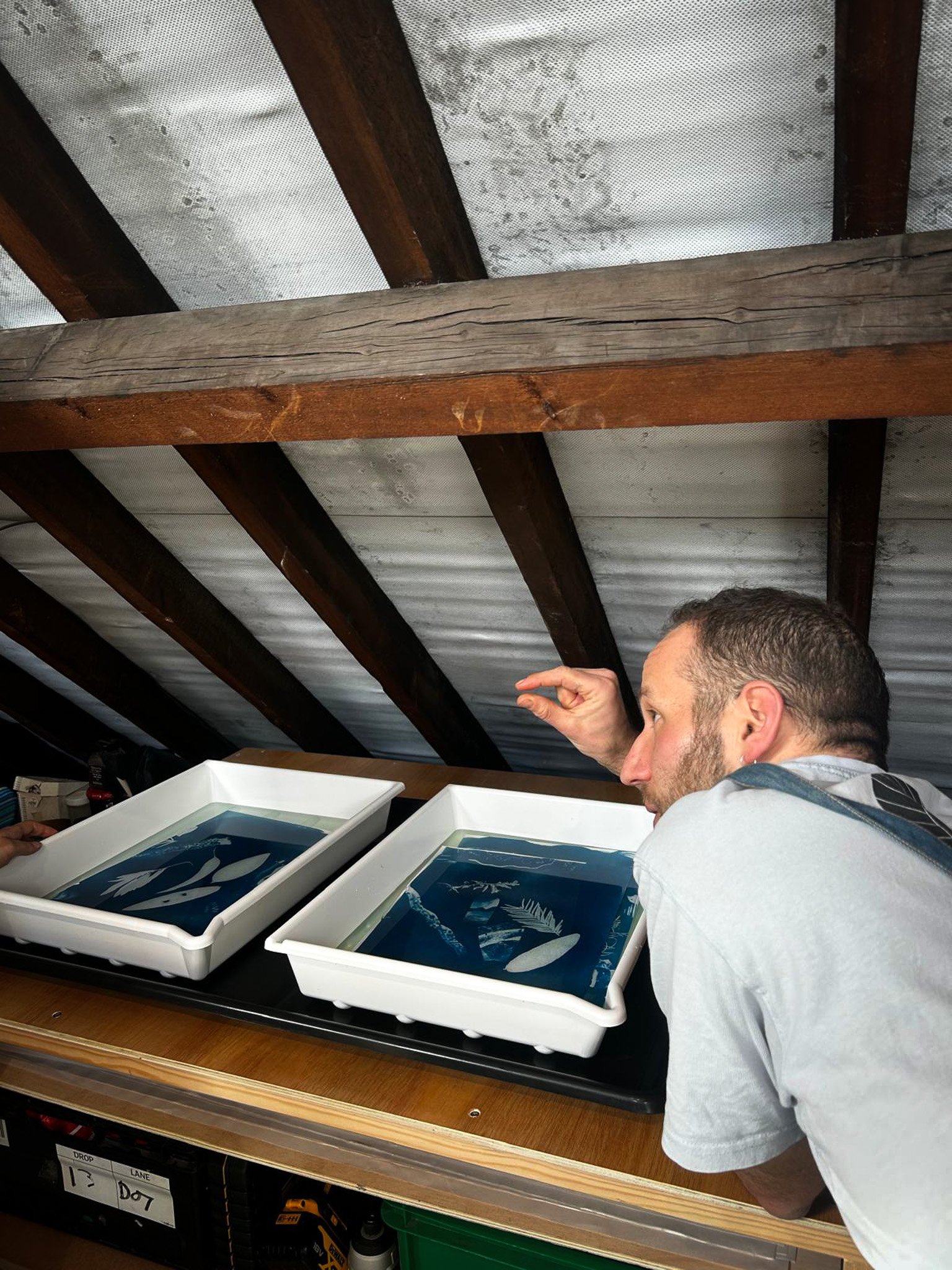 A man with short hair and a beard wearing a gray shirt is looking at white containers filled with dark blue and white abstract designs. The containers are placed on a wooden shelf underneath a slanted ceiling with wooden beams and a white textured surface.