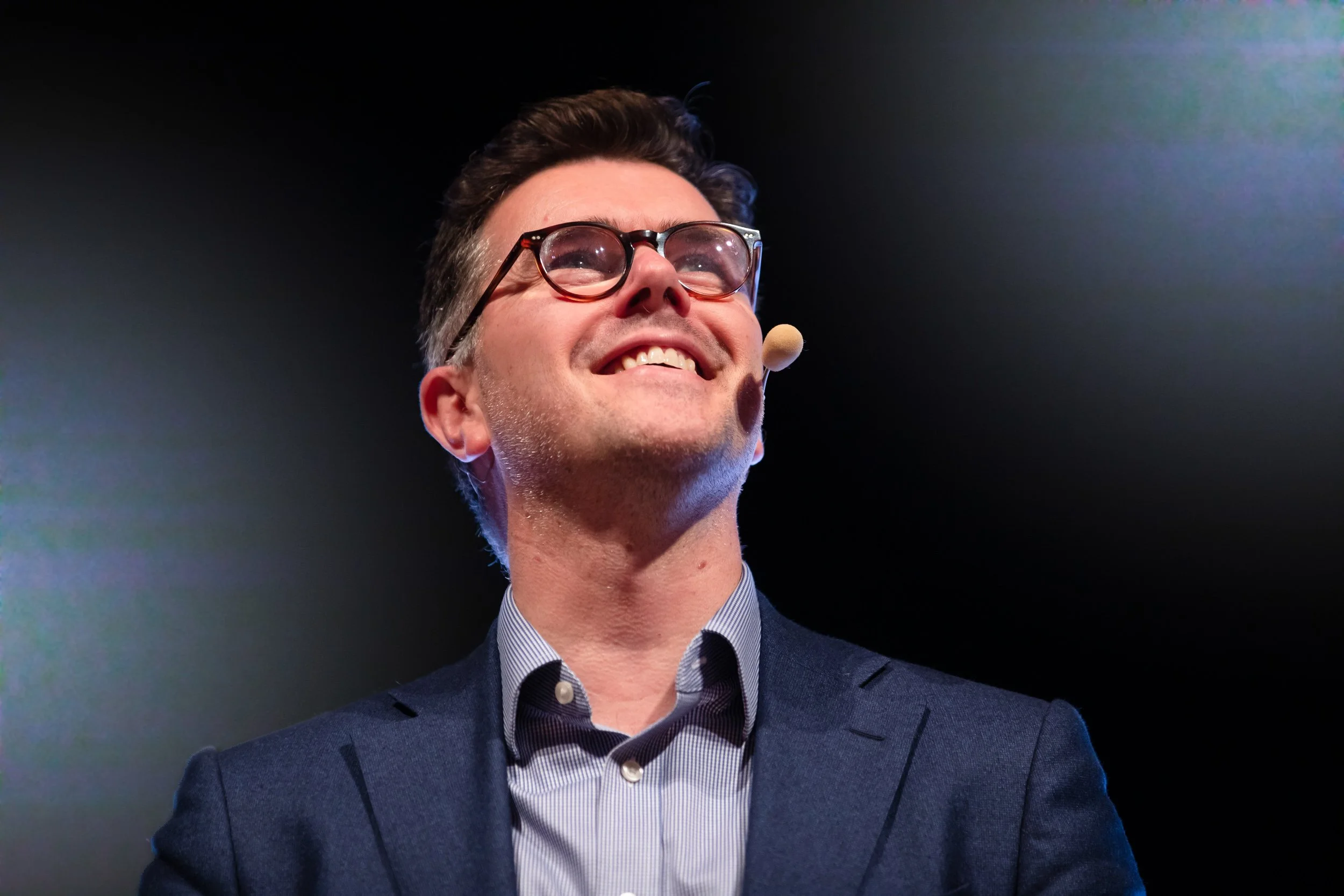 Smiling man with glasses and a microphone headset, wearing a suit, against a dark background.