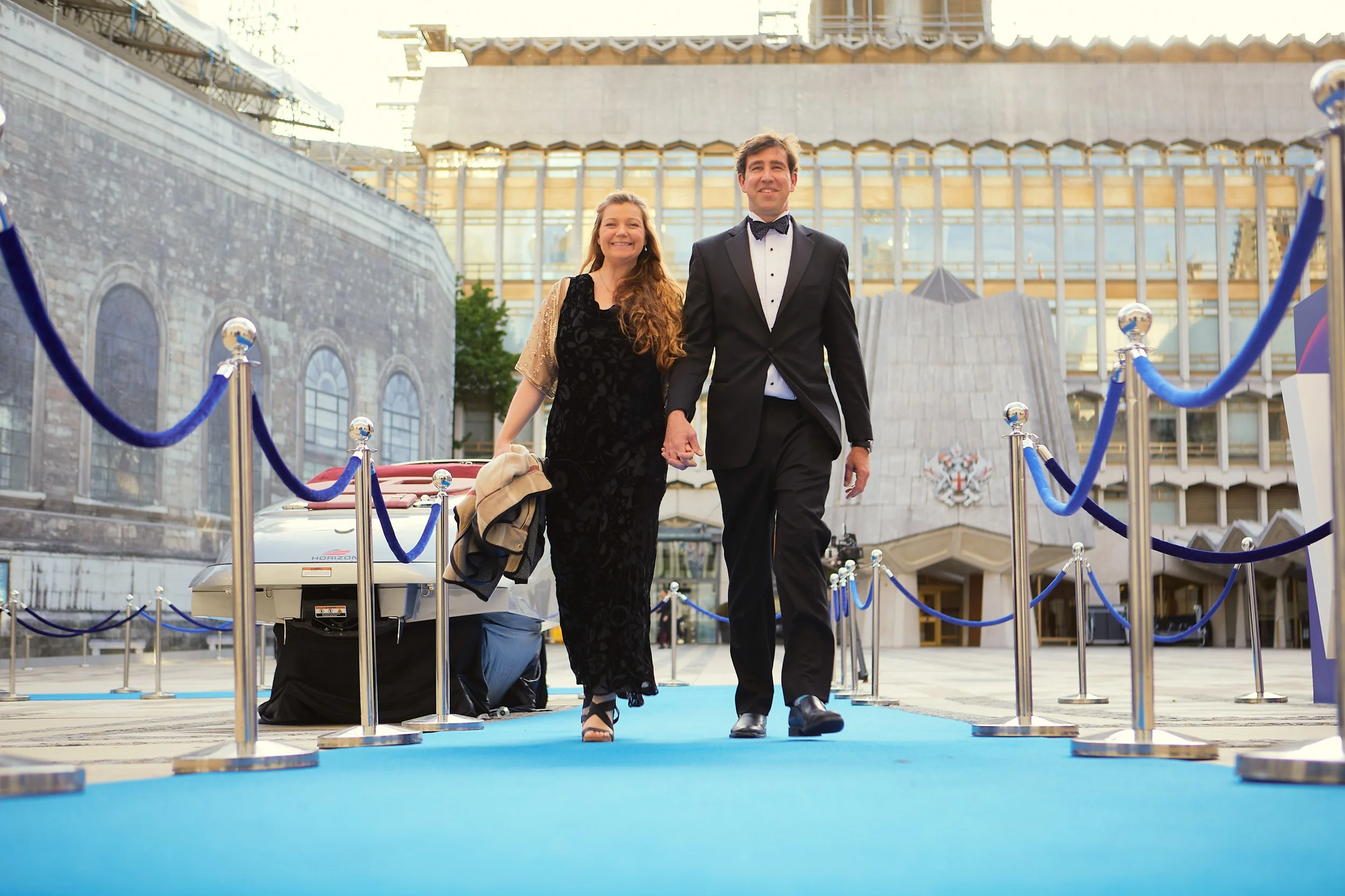 A couple walking on a blue carpet surrounded by stanchions in a formal setting, with a classic car and modern architecture in the background.