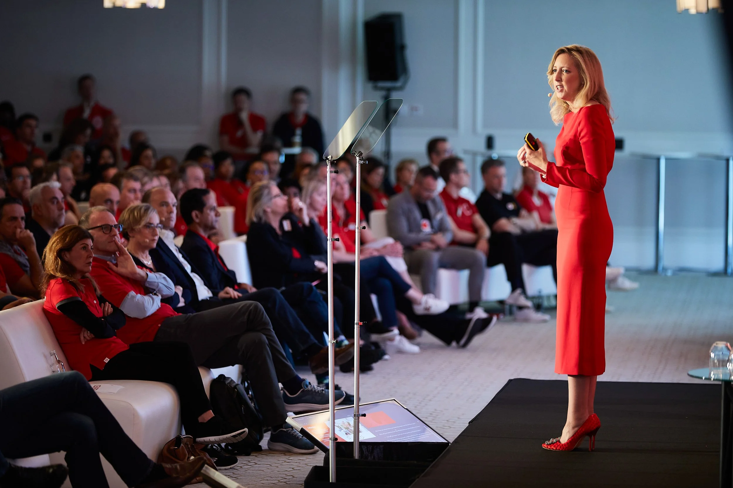 A woman in a red dress speaks on stage at a conference, facing an audience seated in rows. A teleprompter and microphone are visible. The audience is attentive, with some members wearing red polo shirts.