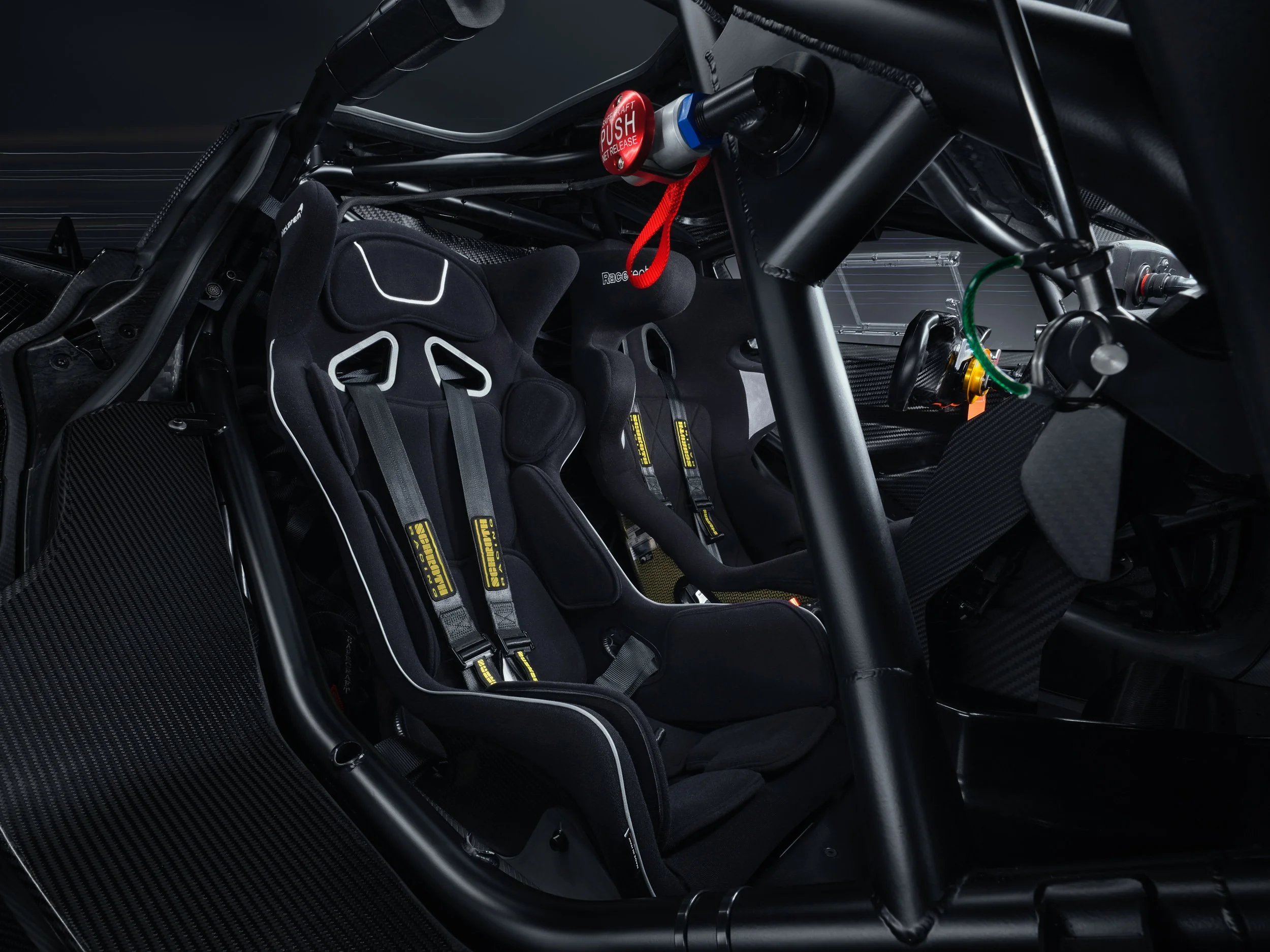 Interior of a high-performance racing car, featuring a black bucket seat with safety harnesses, a roll cage, and carbon fiber components.