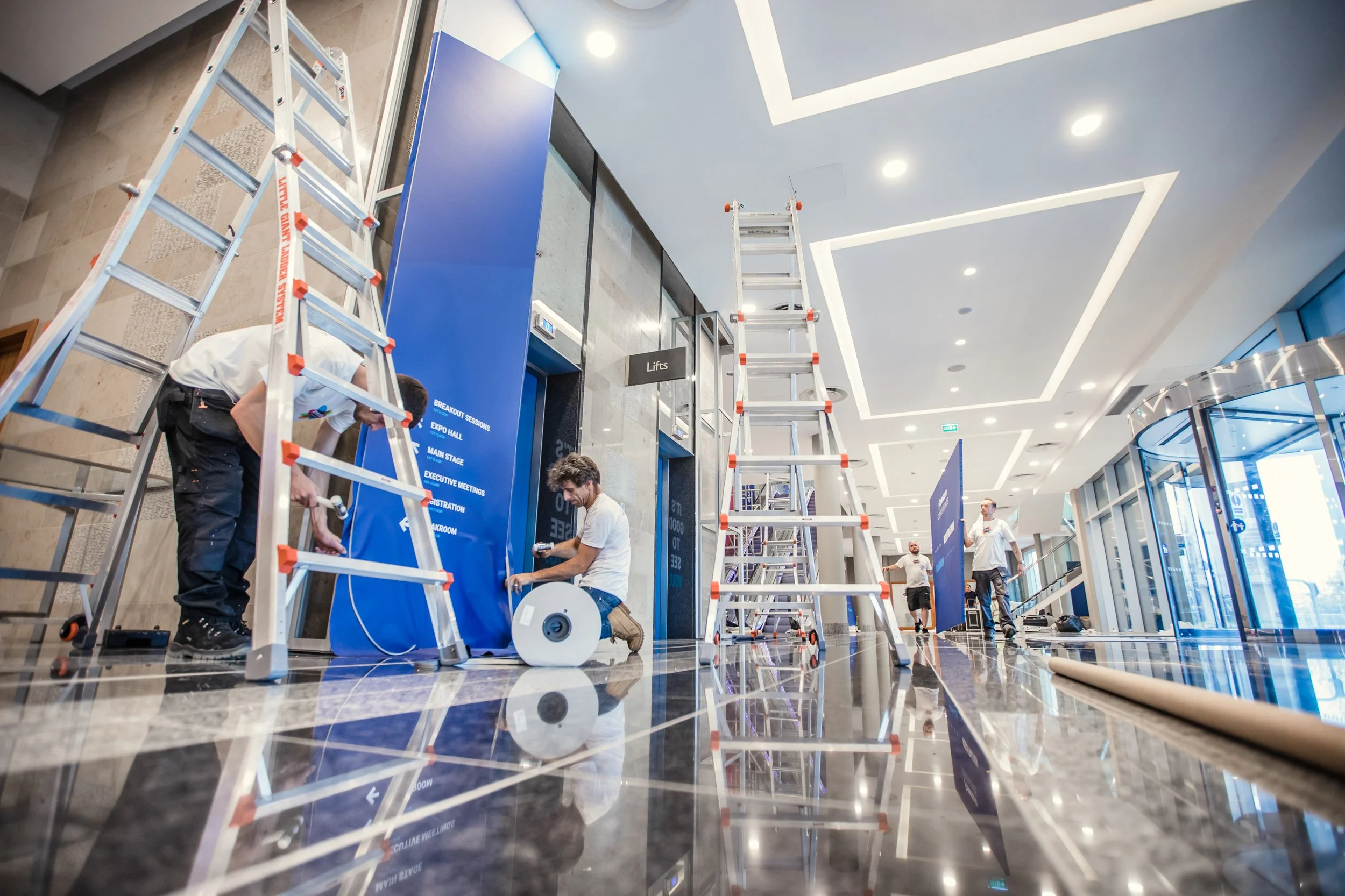 Workers setting up a conference area with ladders, blue panels, and tools in a modern building.