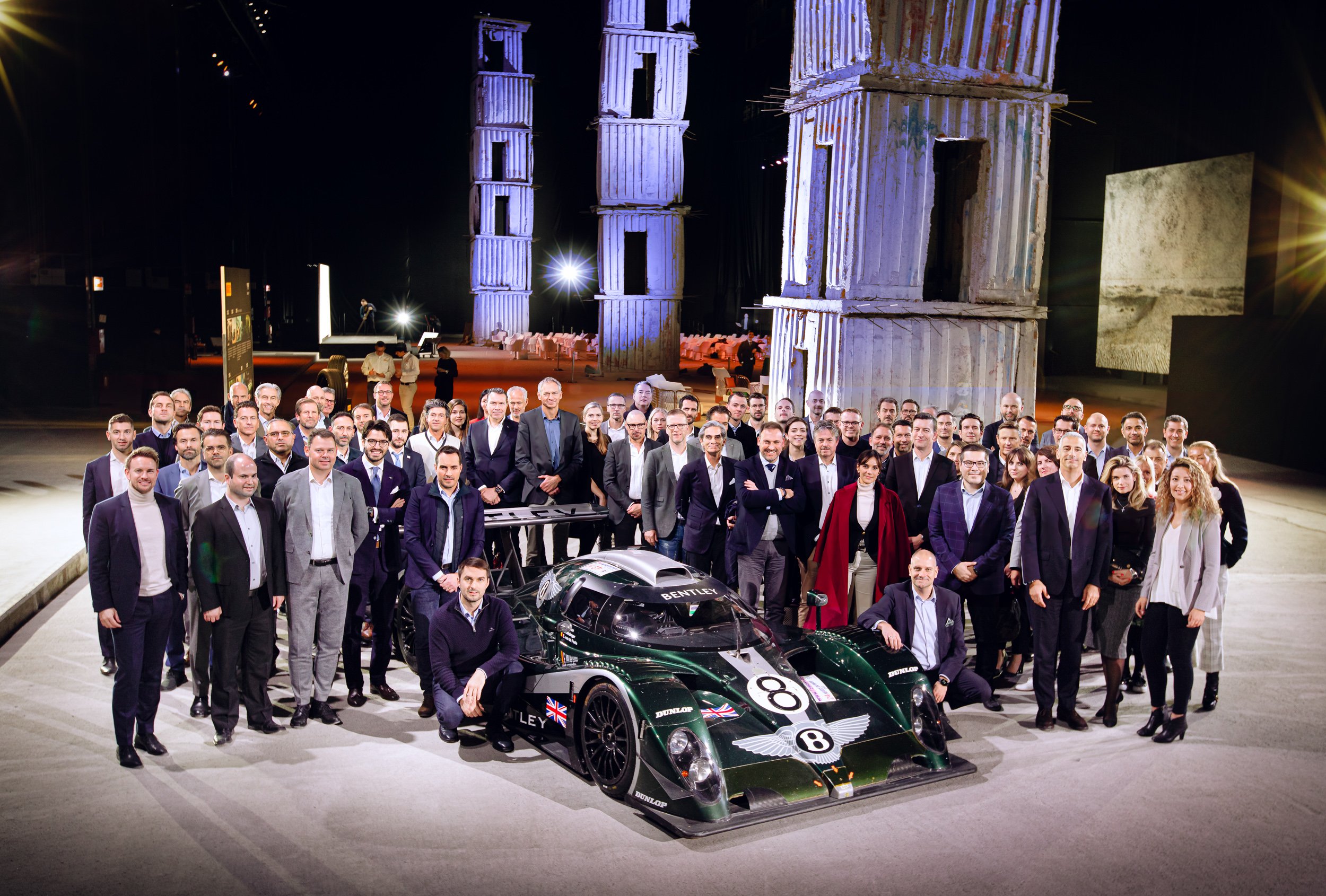 Large group of people posing with a Bentley race car in an industrial setting.