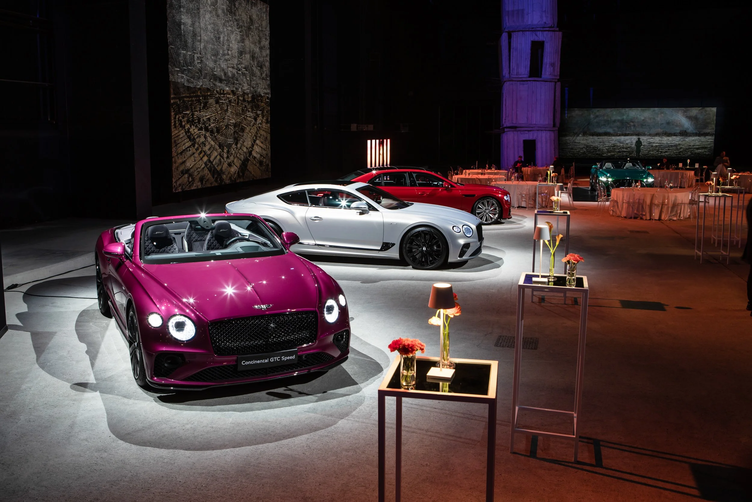 Luxury car showroom with multiple Bentley models on display, including a red convertible, white coupe, and red sedan, set against an elegant event backdrop with flowers and dramatic lighting.