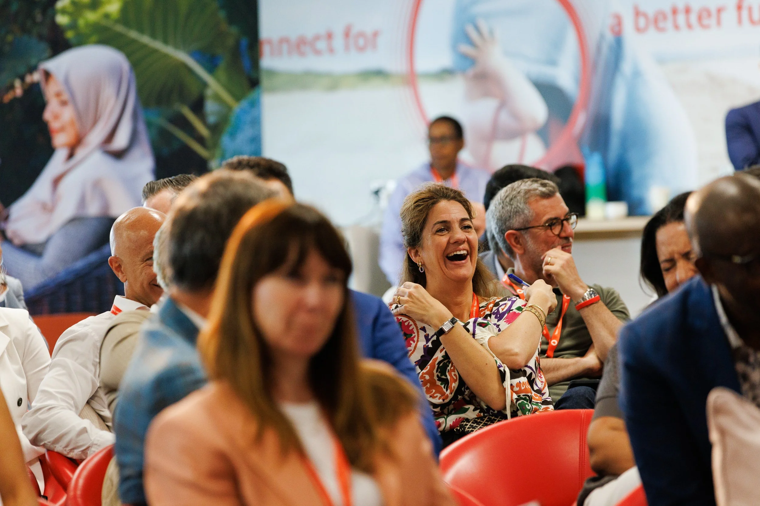Audience seated indoors at an event, with people laughing and engaging, a poster and plants in the background.