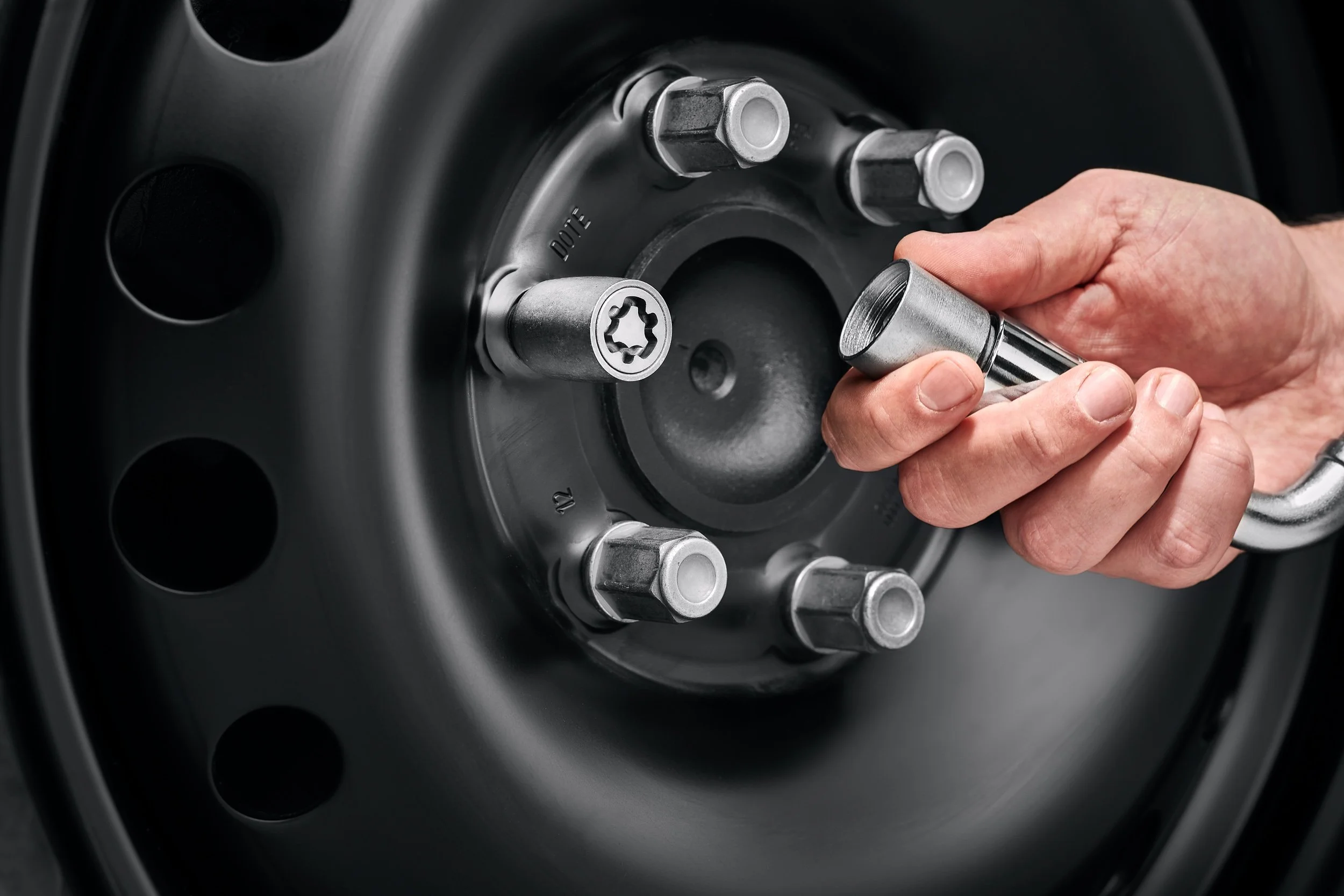 Close-up of a hand holding a socket wrench near a car wheel lug nut with a lock. The wheel is black with metallic lug nuts.