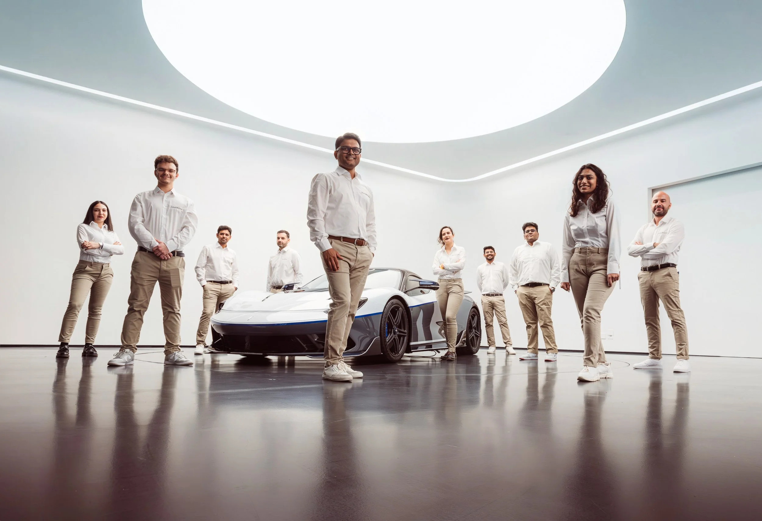 A group of people in white shirts and beige pants standing around a modern sports car in a well-lit room.