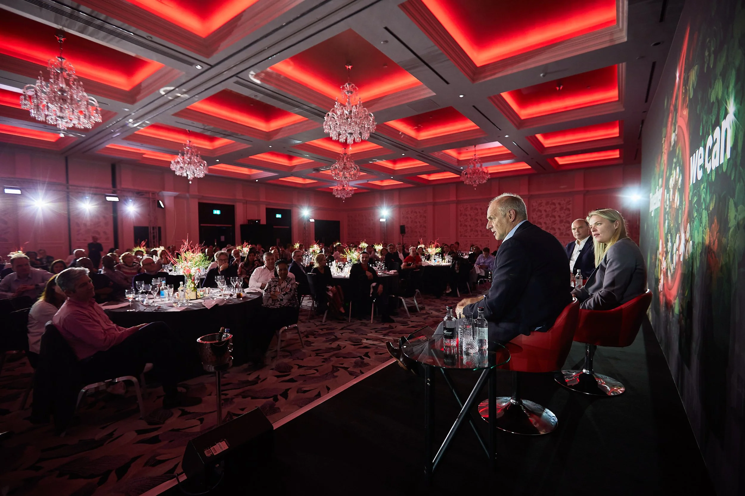 Business conference in a ballroom with red ceiling lights, three panelists seated on stage, and an audience at round tables with floral centerpieces.