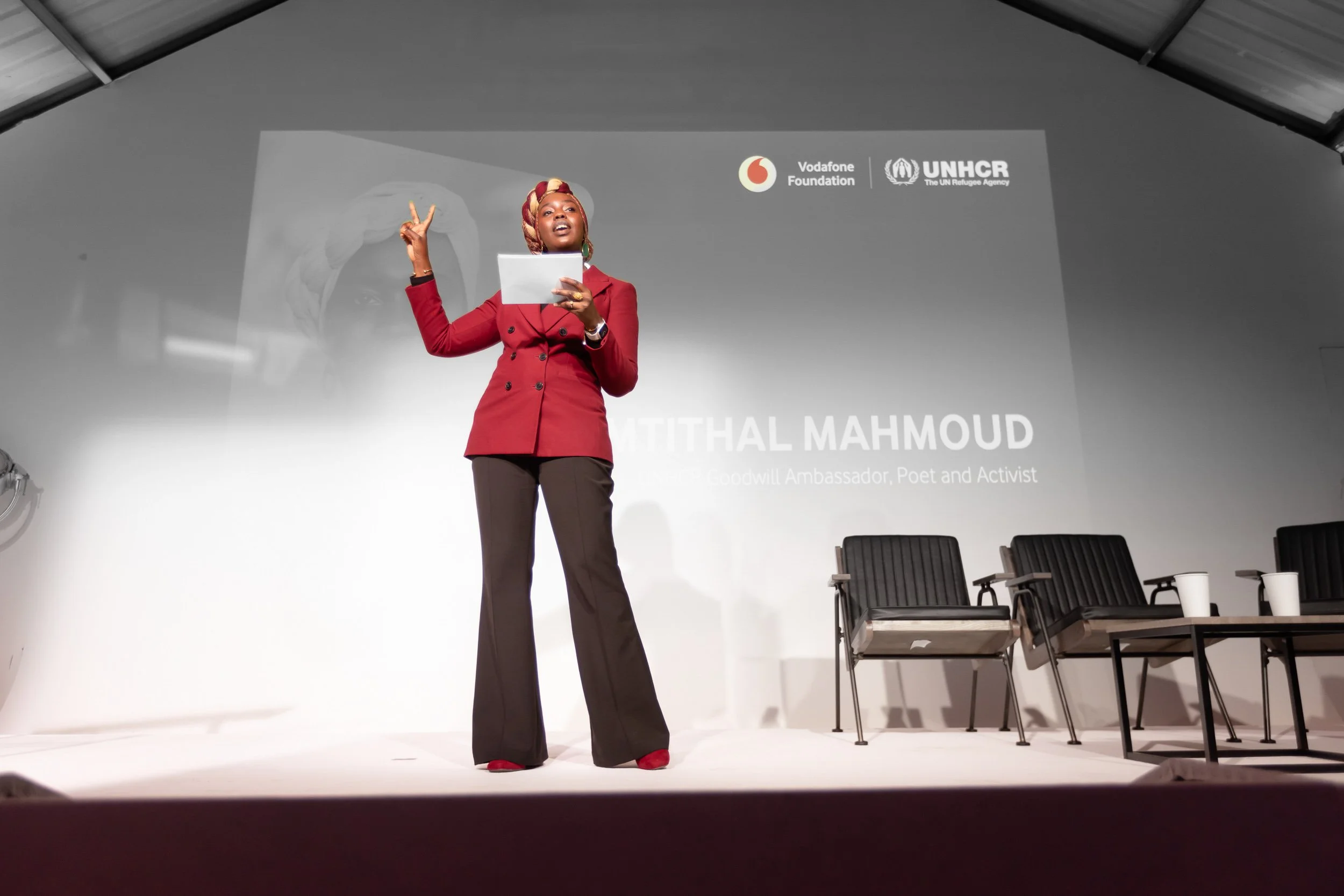 A person in a red outfit speaks on stage at an event. The background shows logos for Vodafone Foundation and UNHCR, along with a name and title indicating the person is a goodwill ambassador, poet, and activist. Empty chairs and tables are visible on