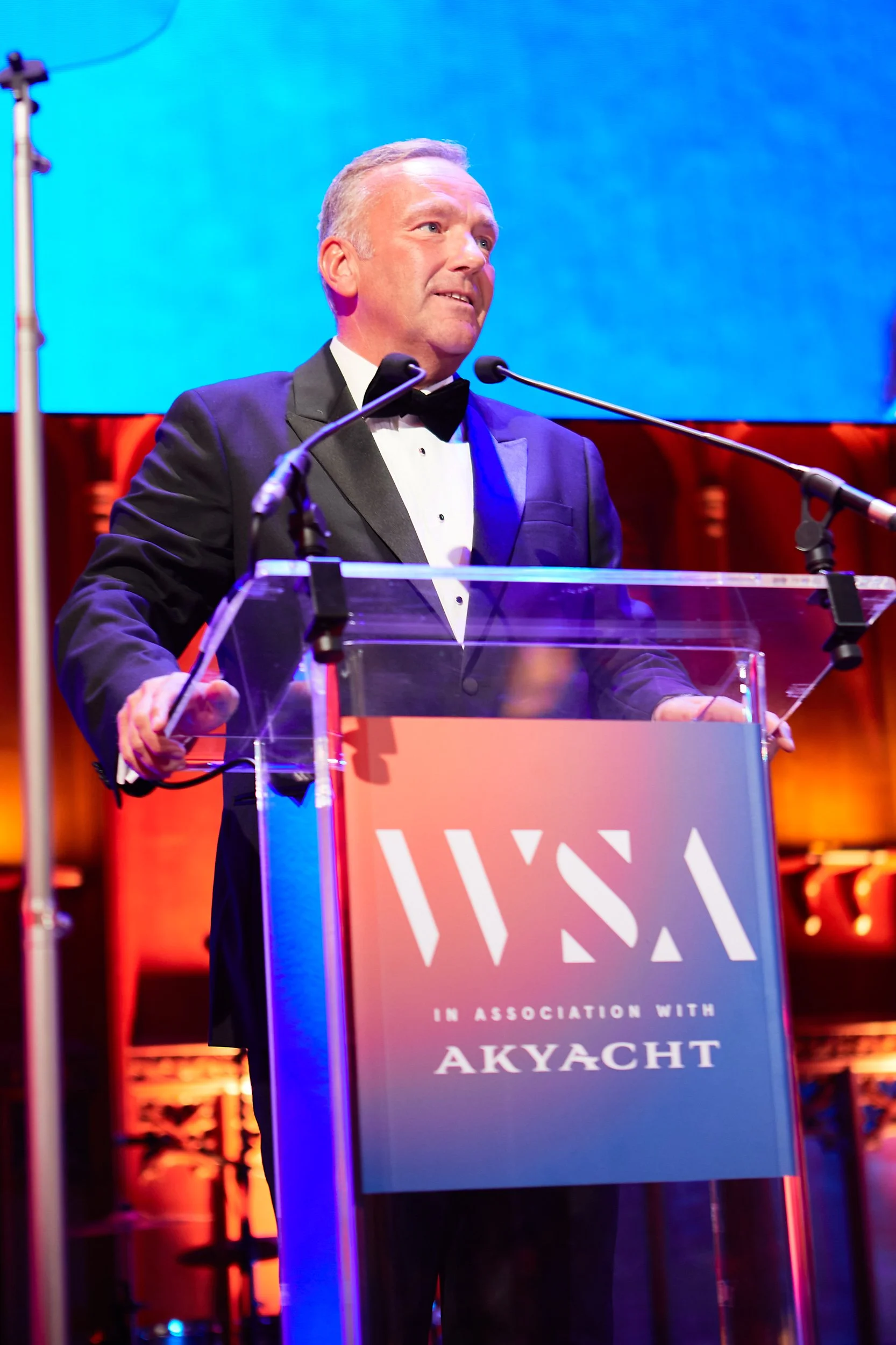 Man in a tuxedo speaking at a podium with "WSA in Association with Akyacht" banner.