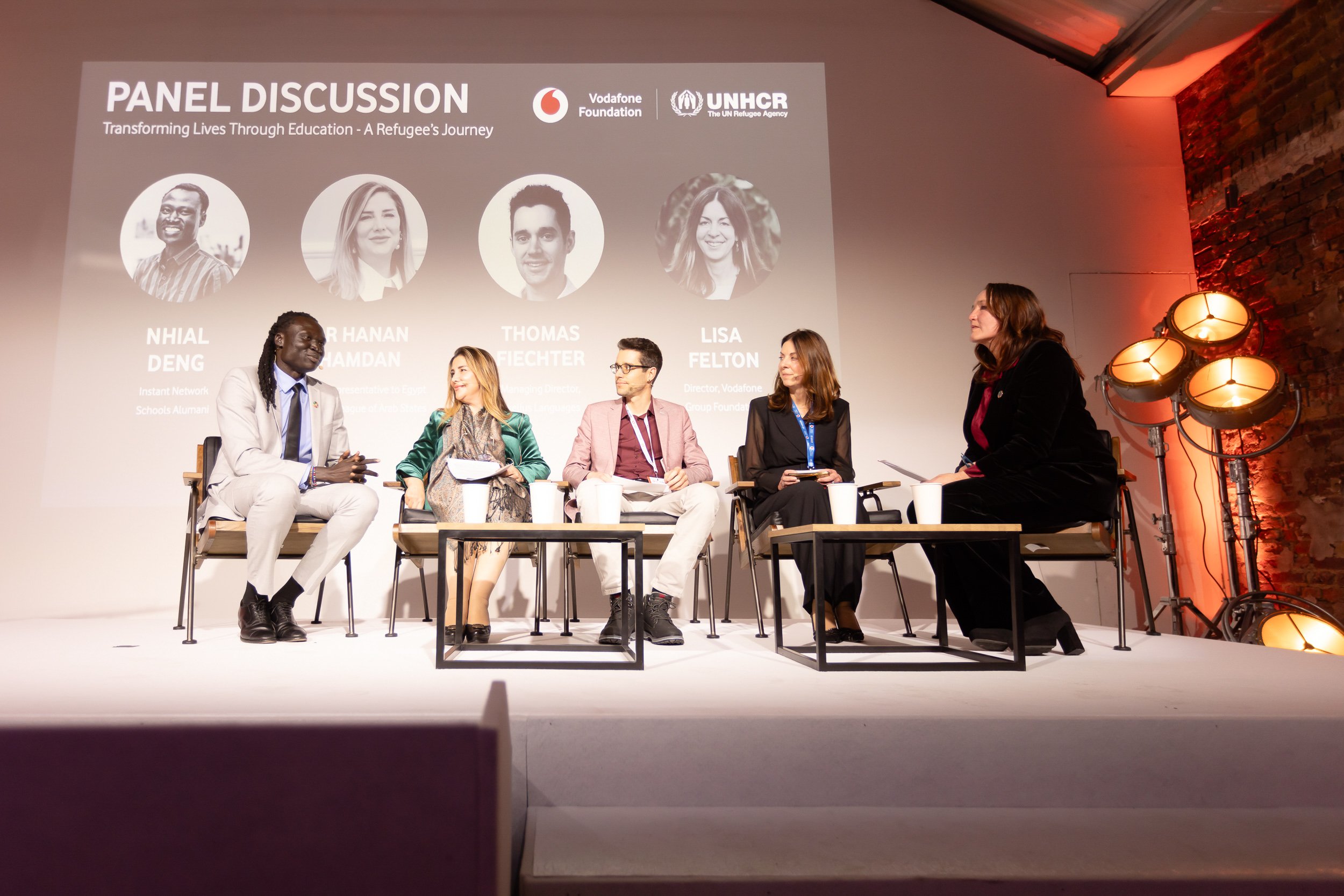 Panel discussion with five participants seated on stage, under a screen displaying event details. The backdrop includes logos for Vodafone Foundation and UNHCR. The participants engage in a conversation about education and refugees.