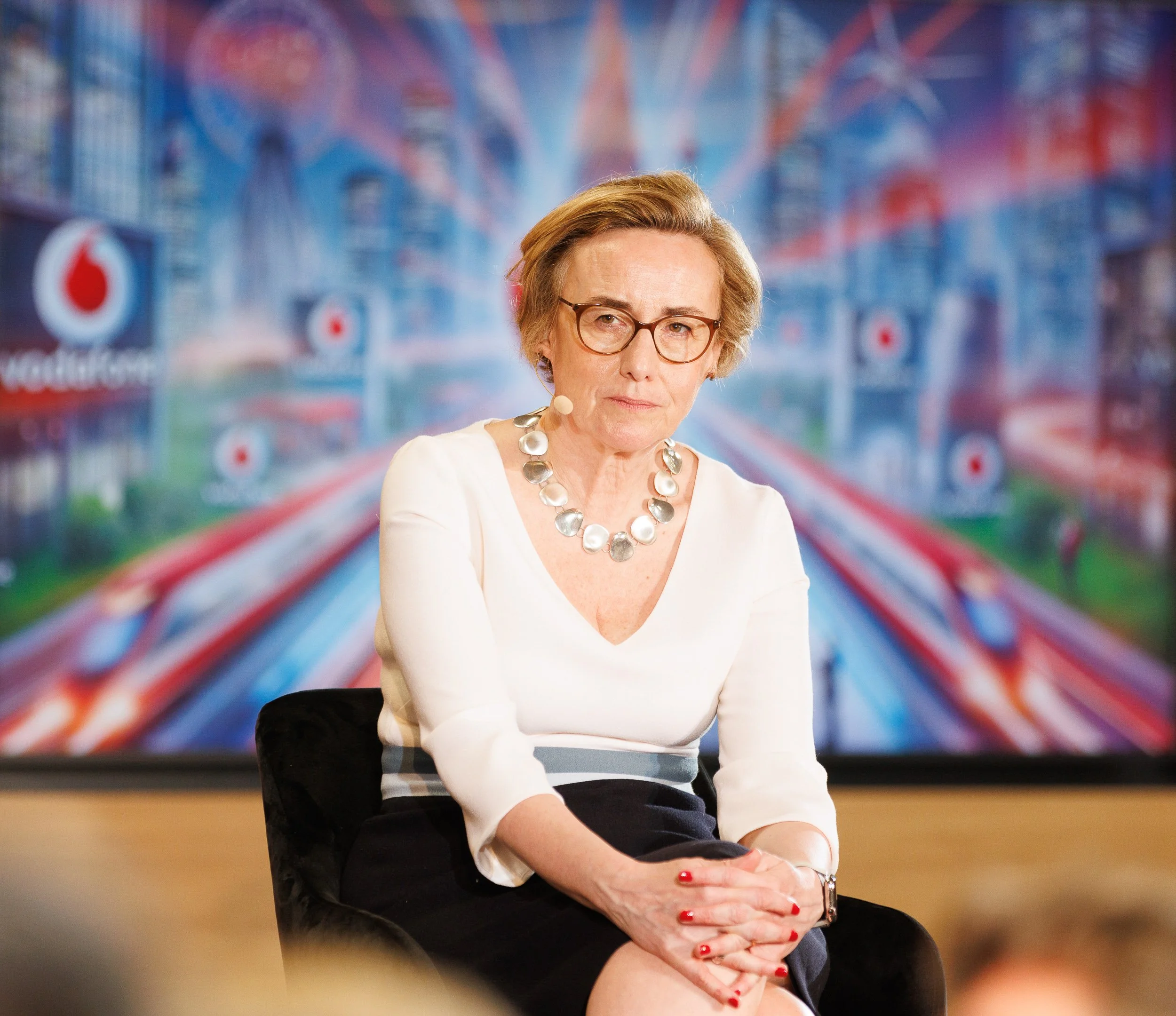 A person sitting in front of a colorful cityscape backdrop with the Vodafone logo visible. They are wearing glasses, a white top, and a silver necklace.