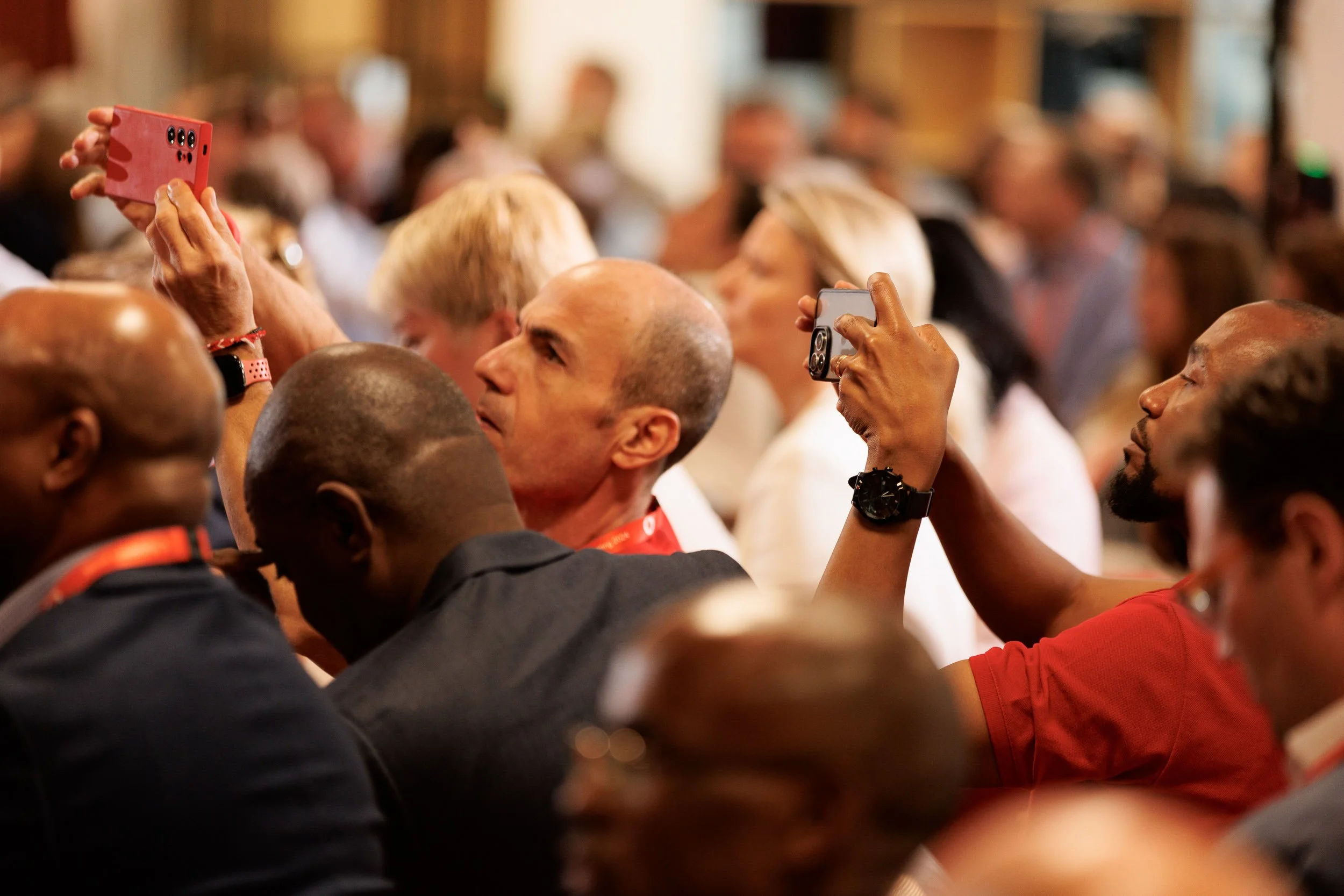 Audience members taking photos with smartphones at an indoor event, focusing on a diverse group of people engaged and attentive.