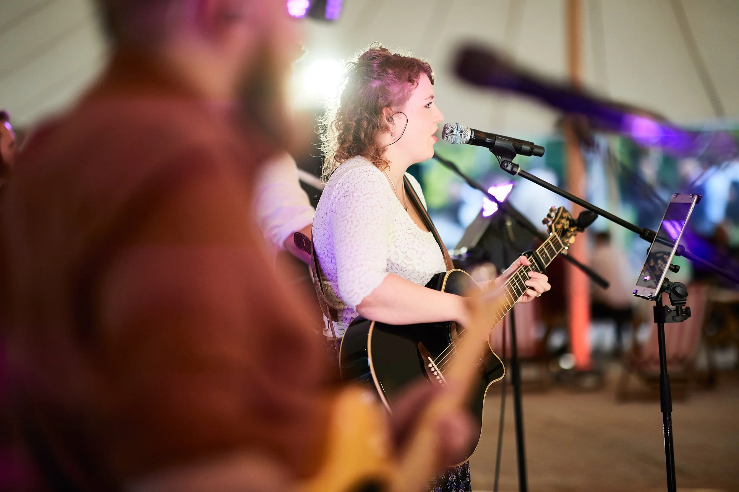 Woman playing guitar and singing into a microphone on stage, with another musician in the foreground, during a live performance.