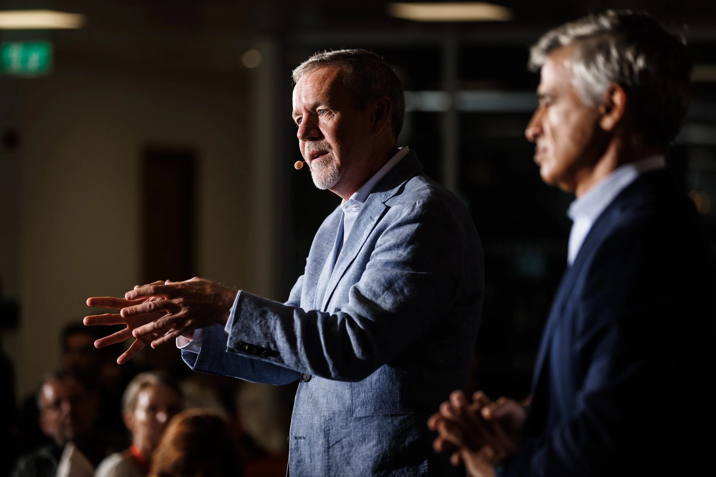 Two men in formal suits speaking at an event, one with a microphone headset, audience in the background.