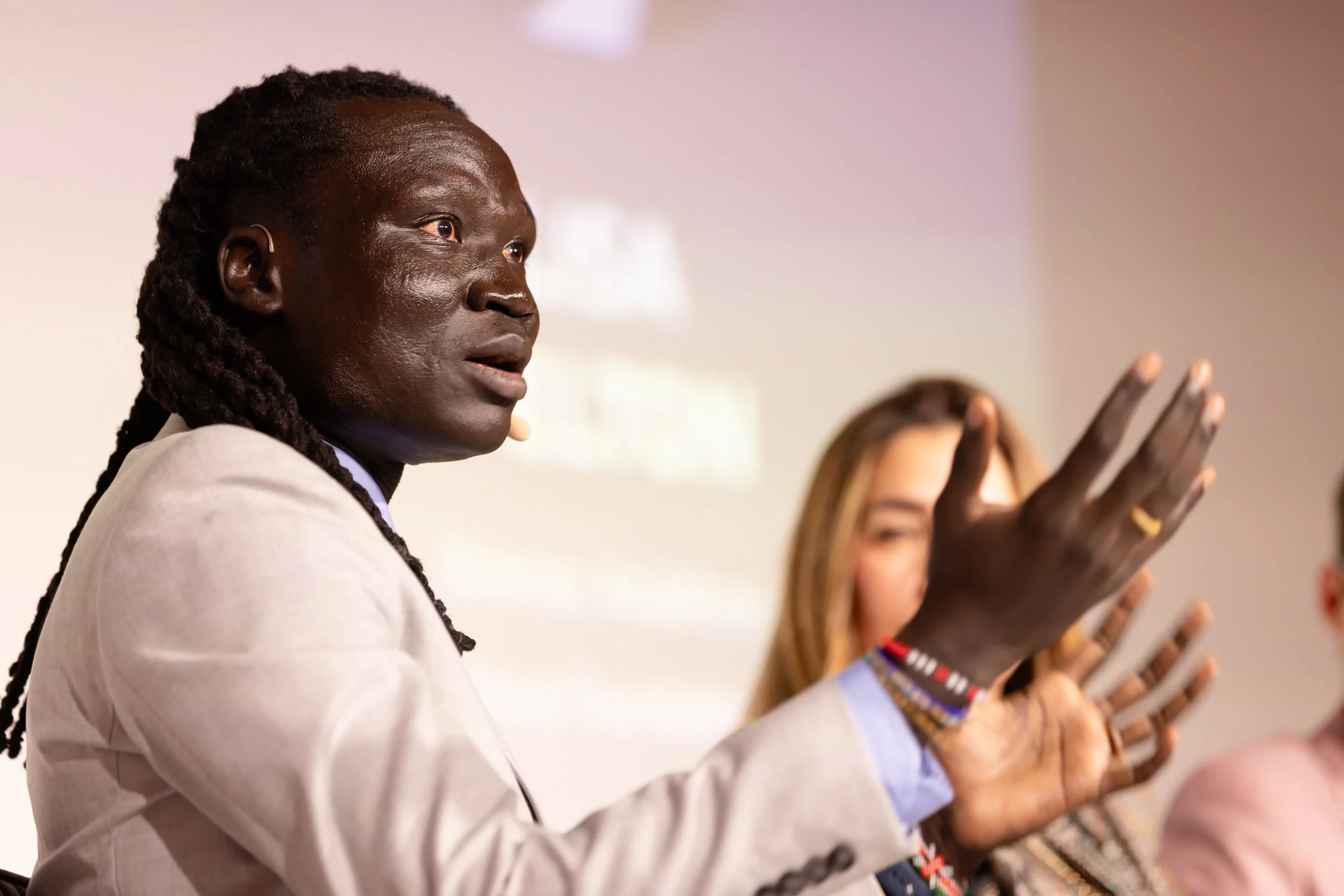 A person in a light gray suit speaking animatedly at a panel discussion, gesturing with their hands. Other blurred panelists are in the background. The semi-blurred background suggests a conference or formal event.