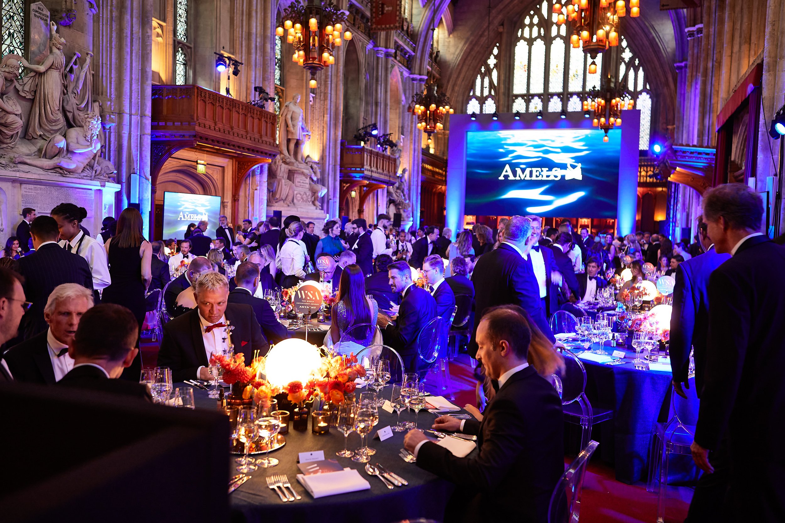 Elegant banquet in a grand hall with chandeliers, sculptures, and large video screen. Guests in formal attire seated at round tables adorned with flowers and candles.
