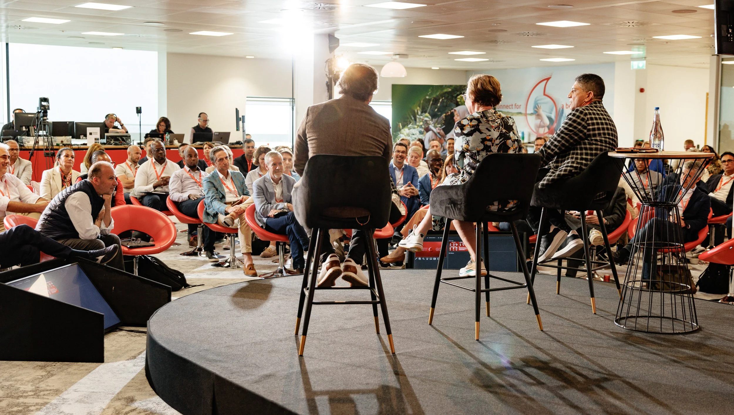 Indoor panel discussion at a conference with three speakers seated on stage chairs facing an audience. Participants are sitting in red chairs, and some are wearing red lanyards. Background shows people working at computers.