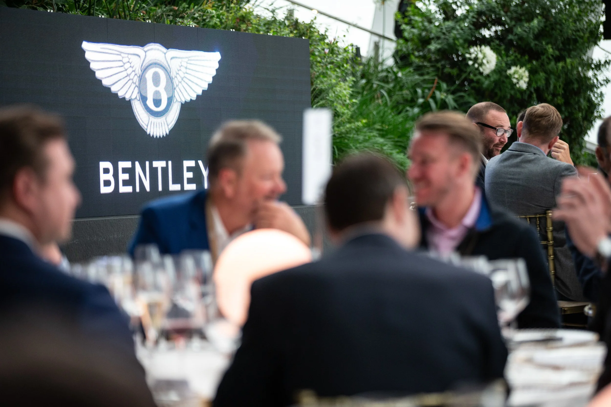People sitting at tables during a Bentley event with logo displayed.