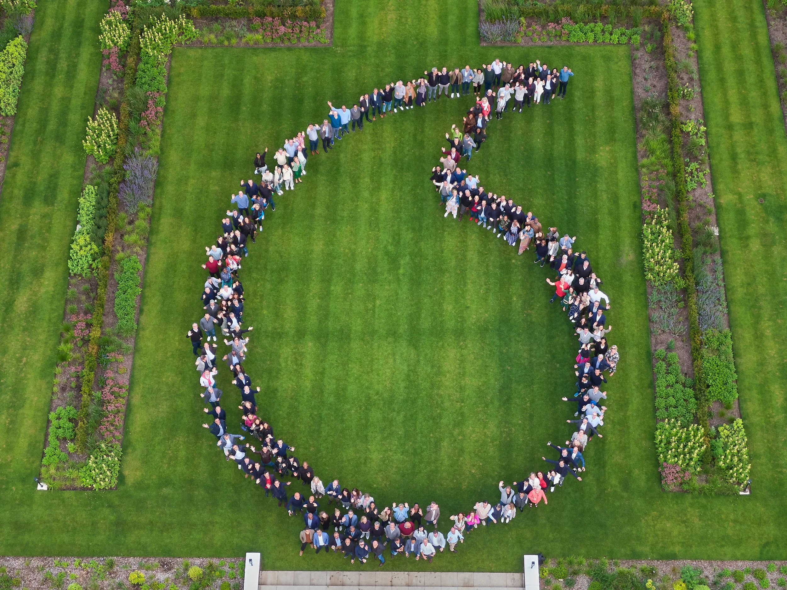 Aerial view of people forming a large comma shape on a grassy lawn surrounded by gardens.