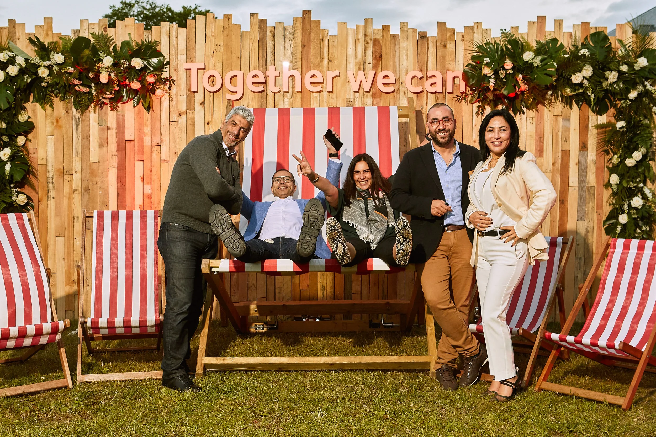 Group of five people posing in front of a large deck chair with a wooden backdrop and flowers, text "Together we can" on top.