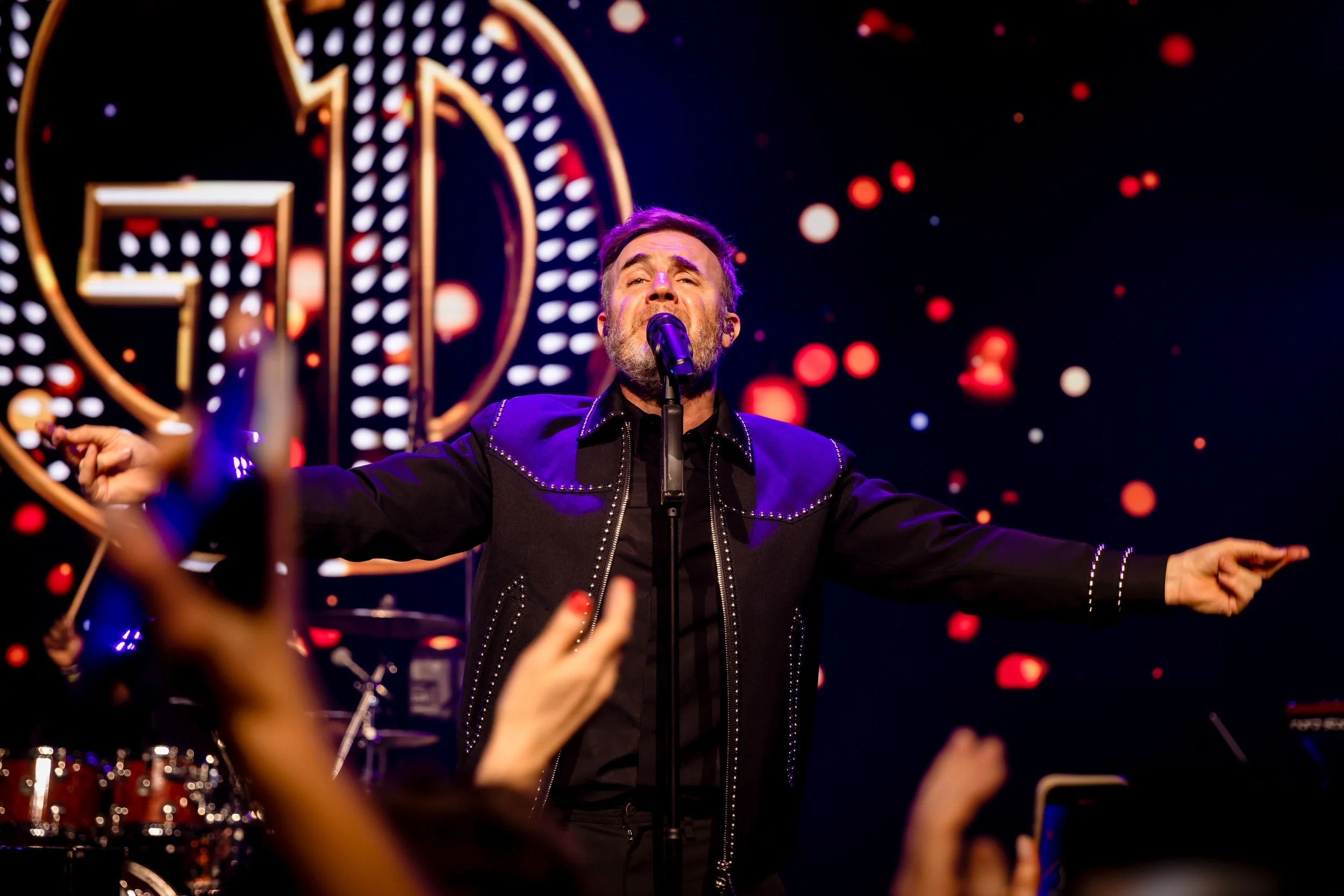 Musician performing on stage with arms outstretched, microphone in front, colorful lights, and audience hands raised.