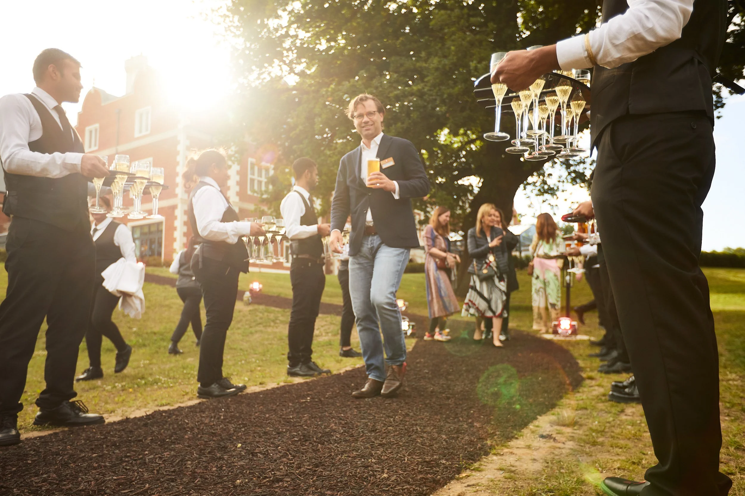 A man walking on a garden path, surrounded by servers in black and white attire holding trays of champagne glasses. In the background, a group of people socializing near a large tree, with a brick building partially visible.