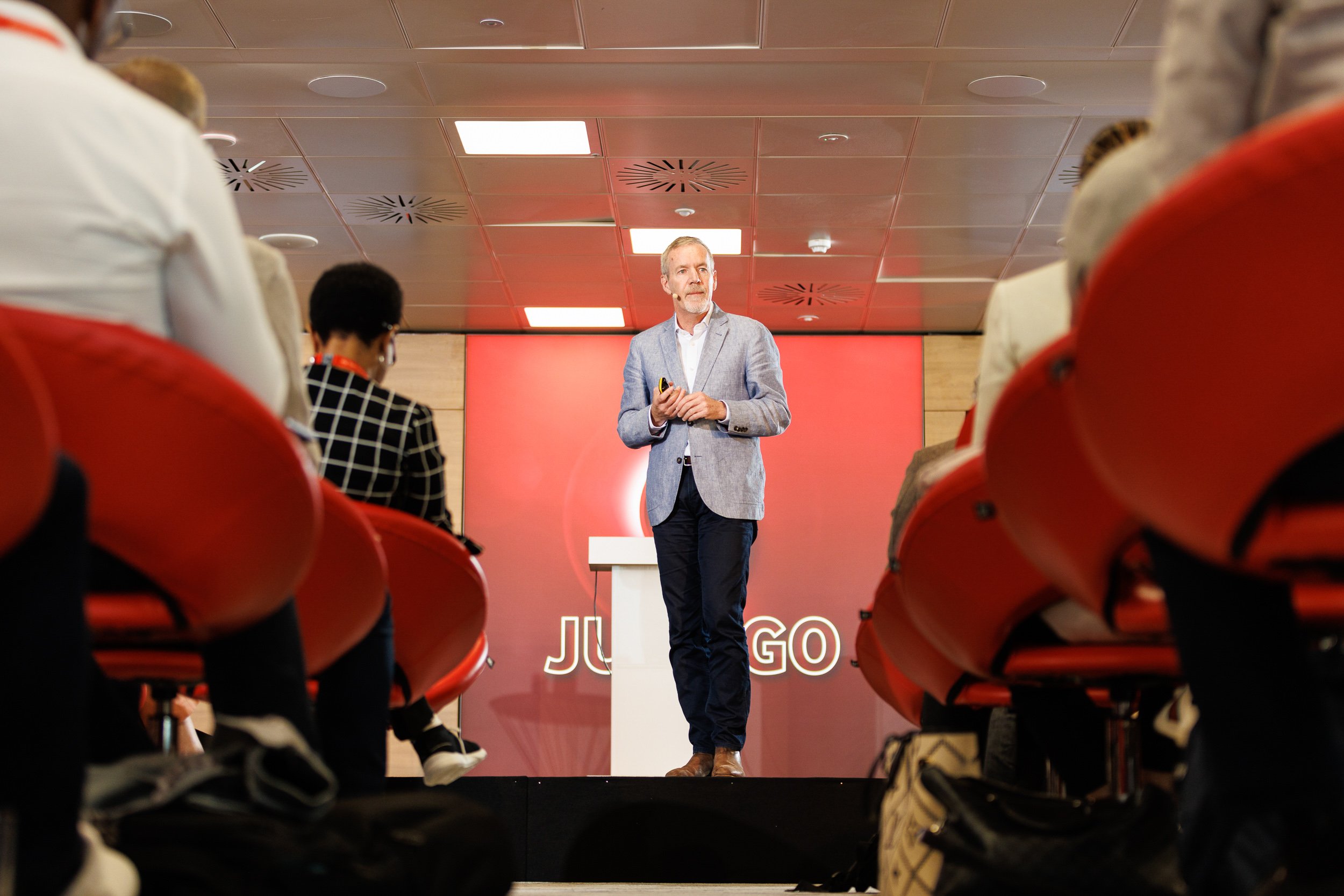 Speaker on stage at a conference, wearing a gray blazer, with audience seated in red chairs