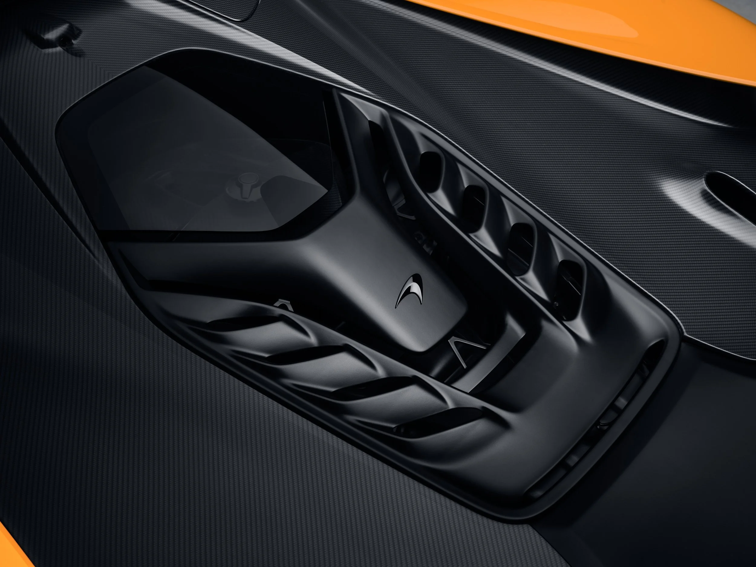 Close-up of a McLaren sports car engine cover with logo, featuring a black carbon fiber texture and orange body detail.