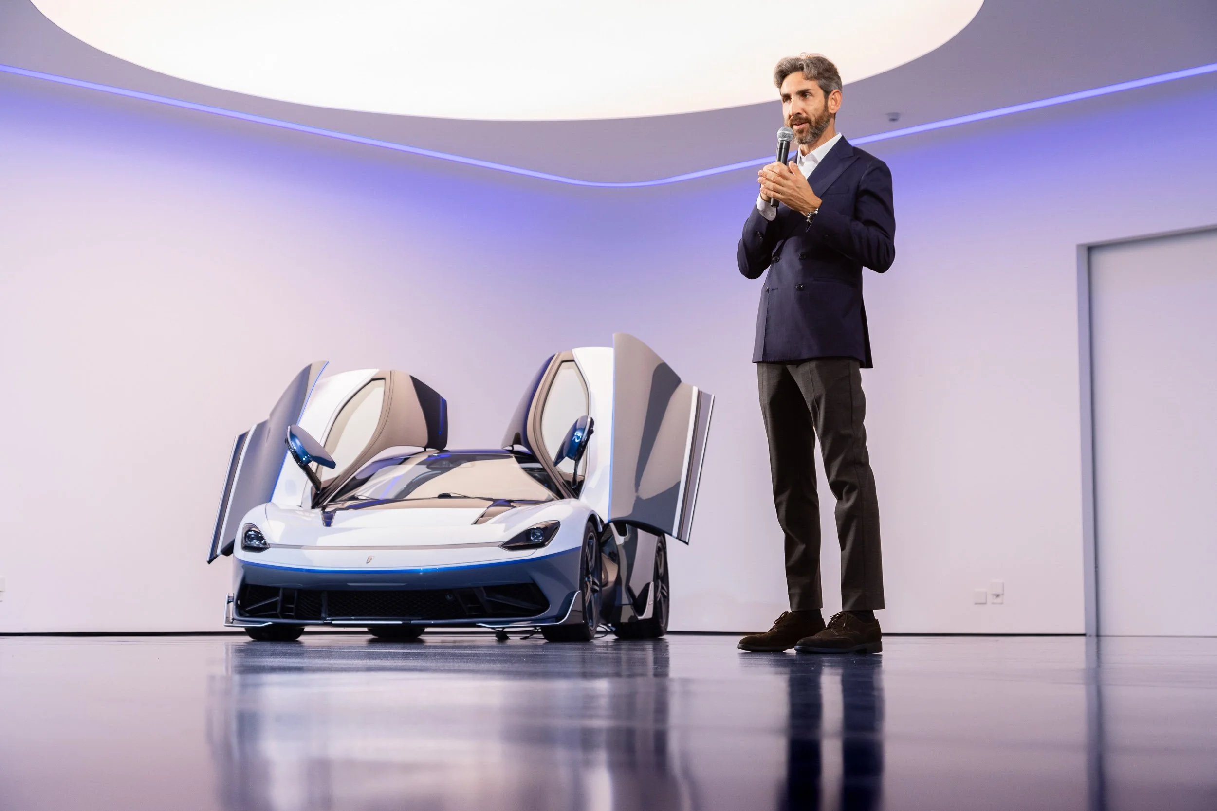 Man standing on stage with a microphone next to a white sports car with open scissor doors.
