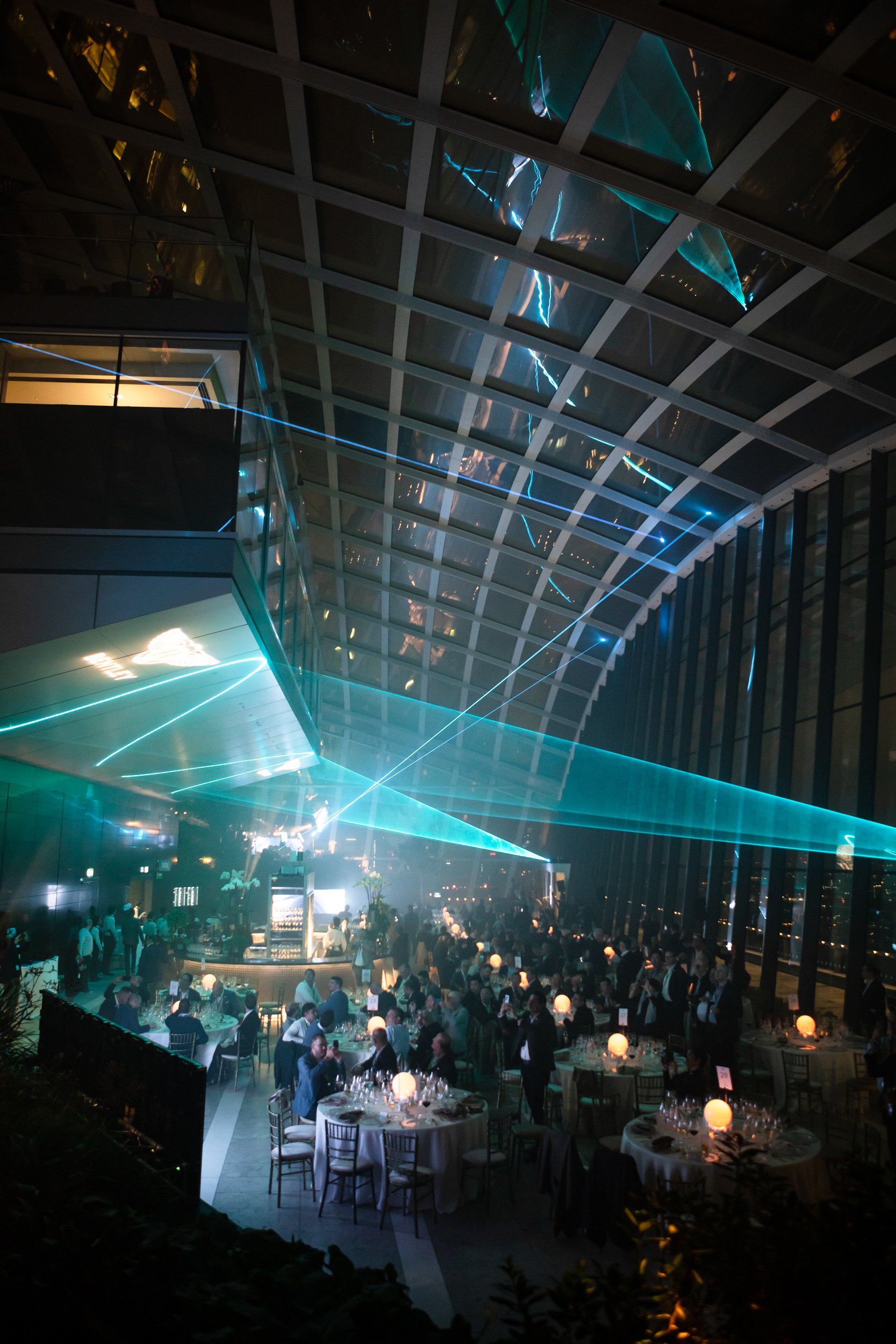 Indoor event with tables, chairs, and attendees under a glass ceiling with laser lights.