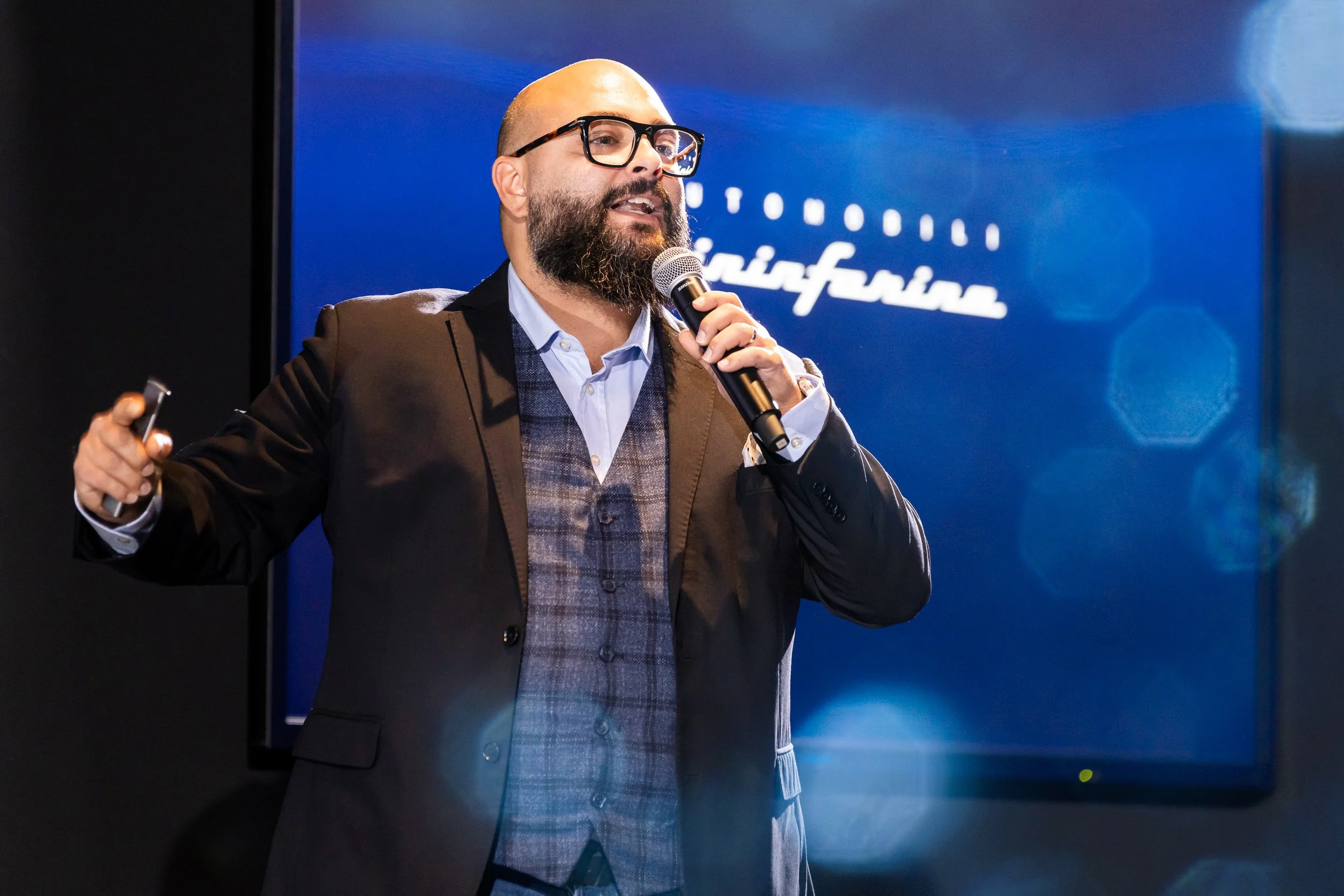 Man giving a presentation with a microphone in hand, standing in front of a screen showing "Automobili Pininfarina" logo.