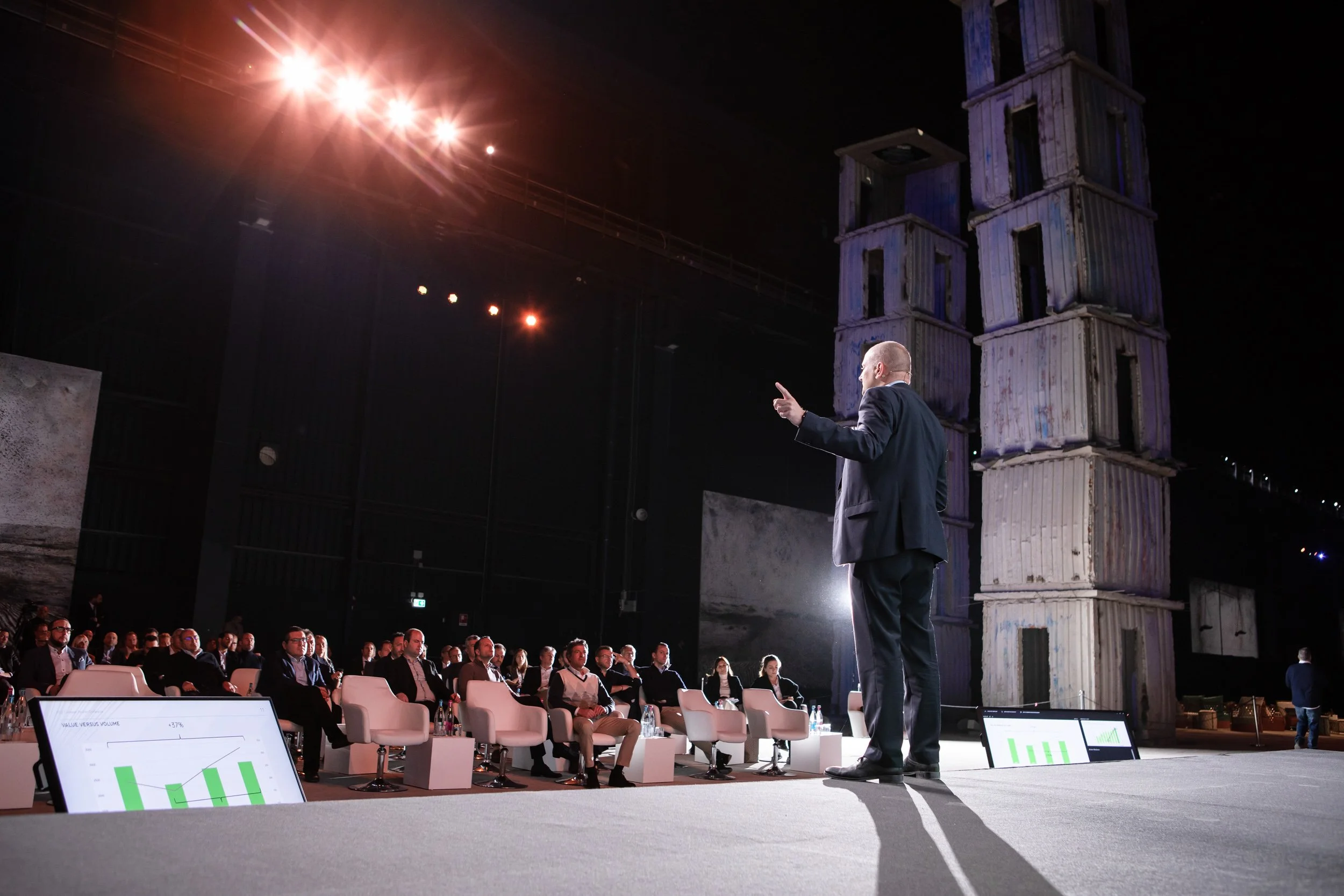 Speaker presenting at a conference with audience seated in front and large screen displaying a graph; tall structure made of stacked containers in background.