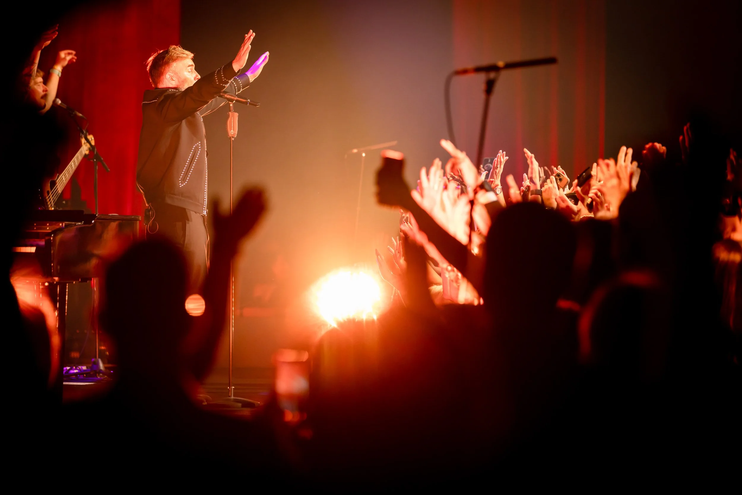 Singer performing on stage with a microphone, arms raised, audience cheering with hands up, illuminated by bright stage lights.