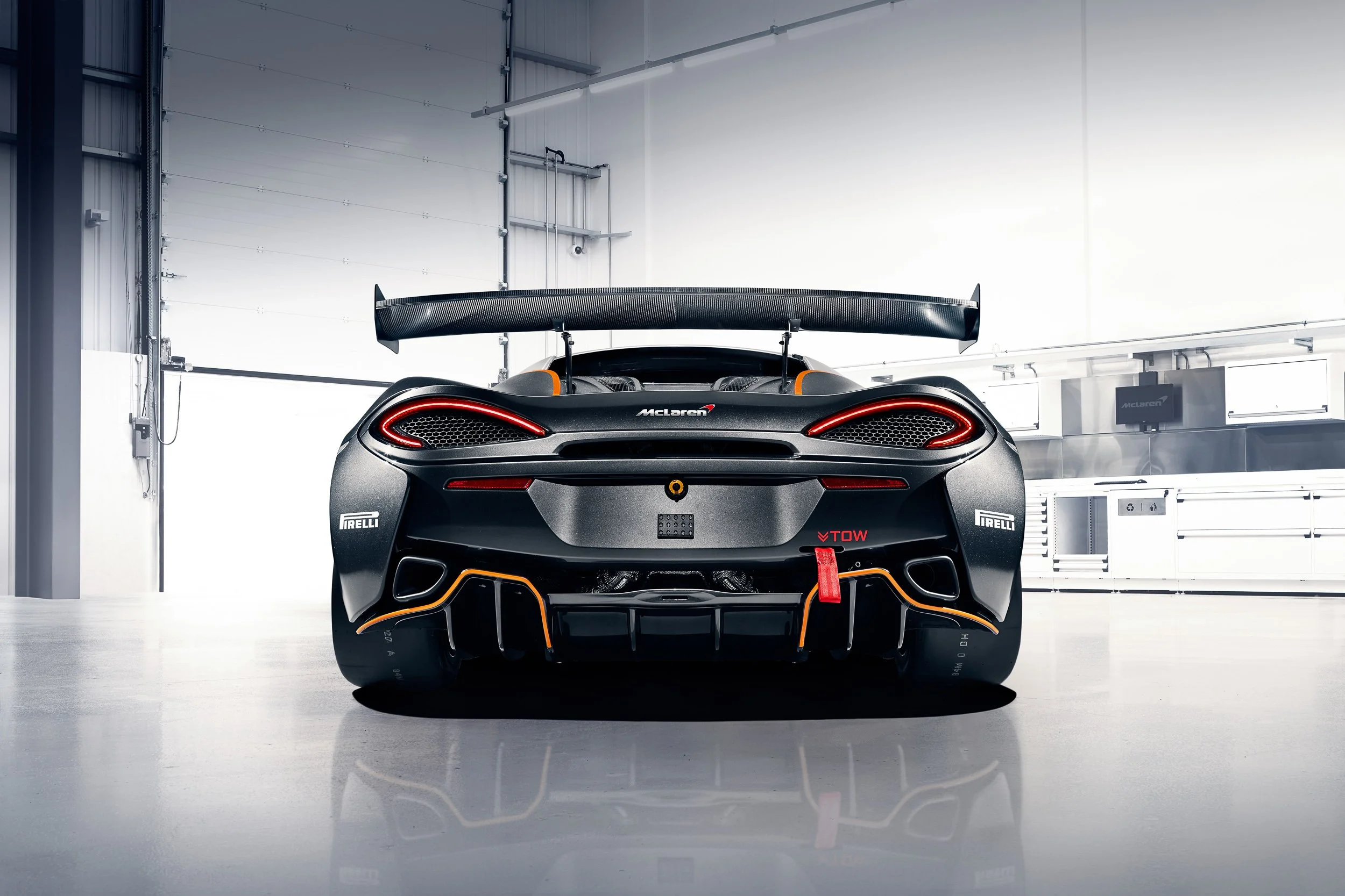 Rear view of a McLaren performance car in a garage setting, featuring a large rear wing, distinctive taillights, and sponsor logos.