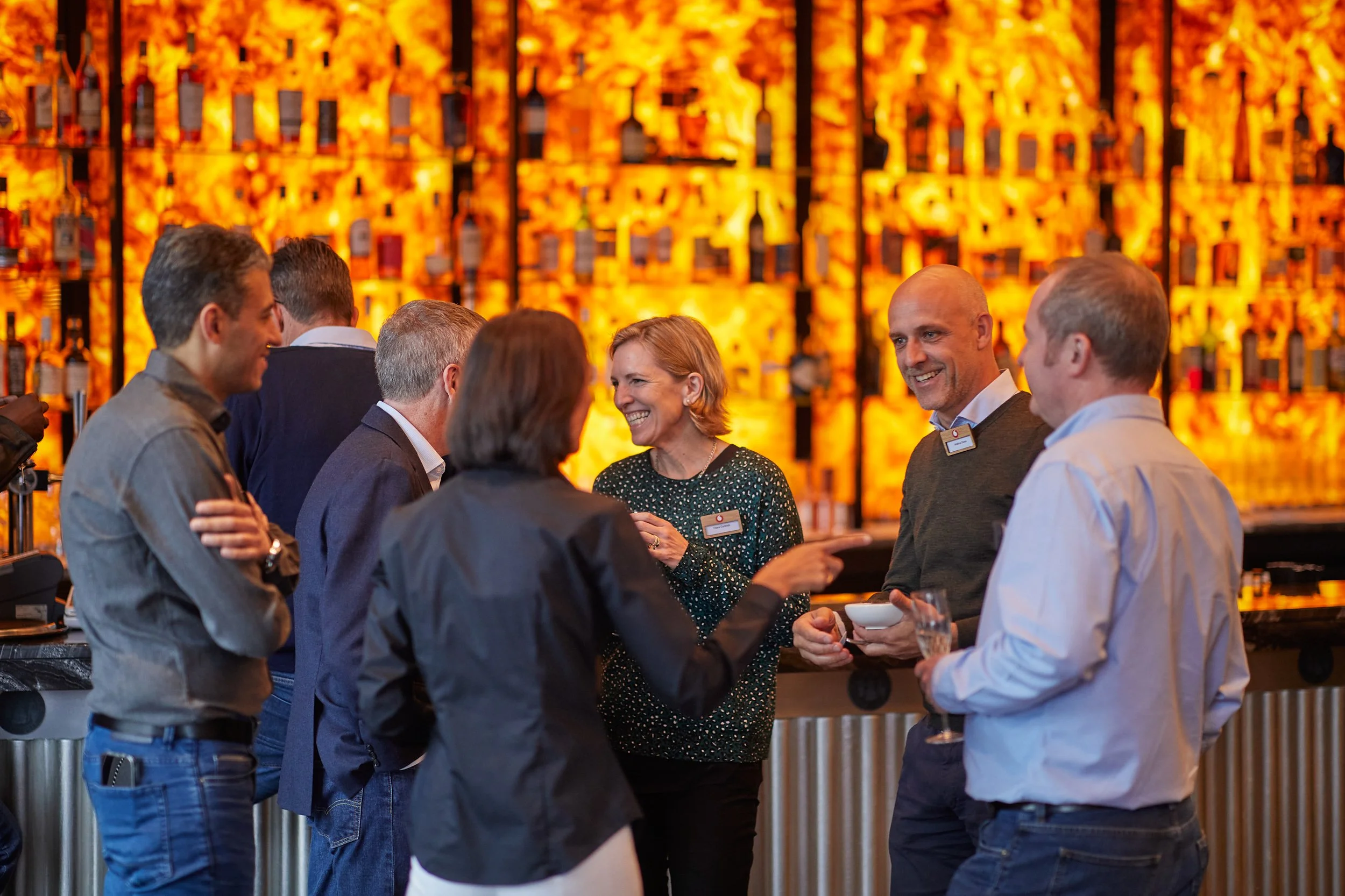 Group of people socializing at a bar with a lit-up wall of bottles.
